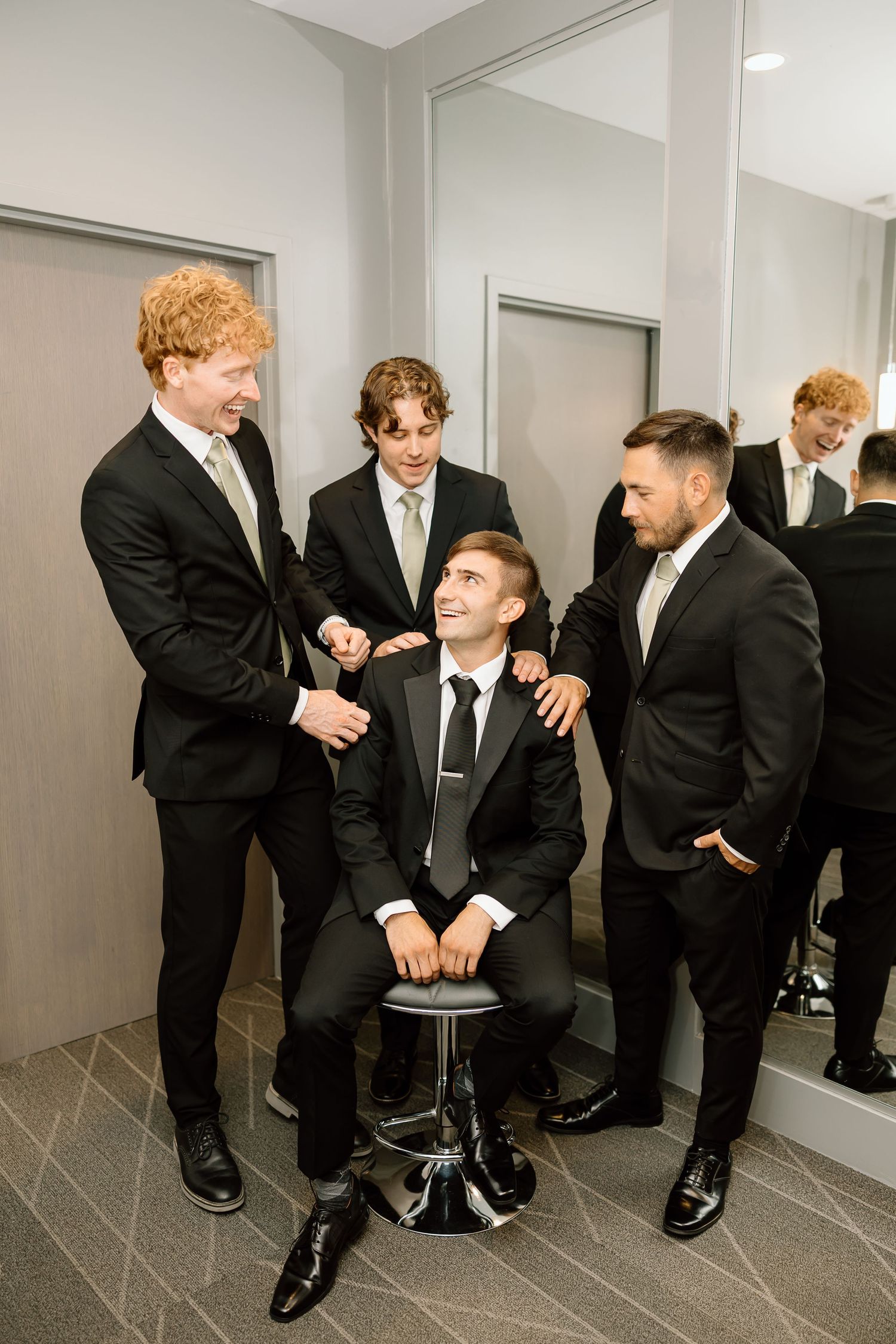 Group in formal black suits playfully interact in a minimalist hallway, creating candid wedding moments.