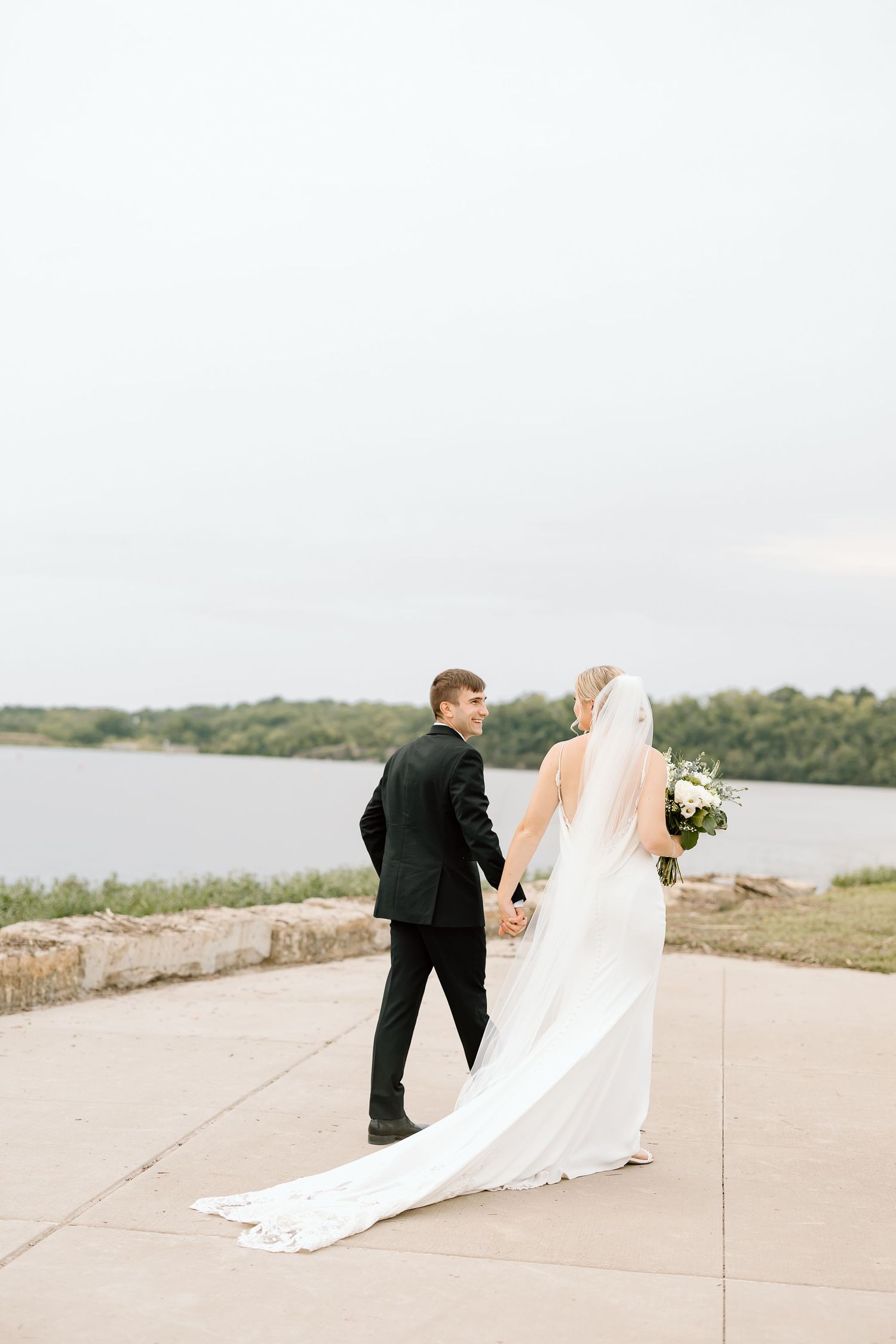 A romantic bride and groom hold hands while walking along a lakeside pathway in a scenic outdoor setting.