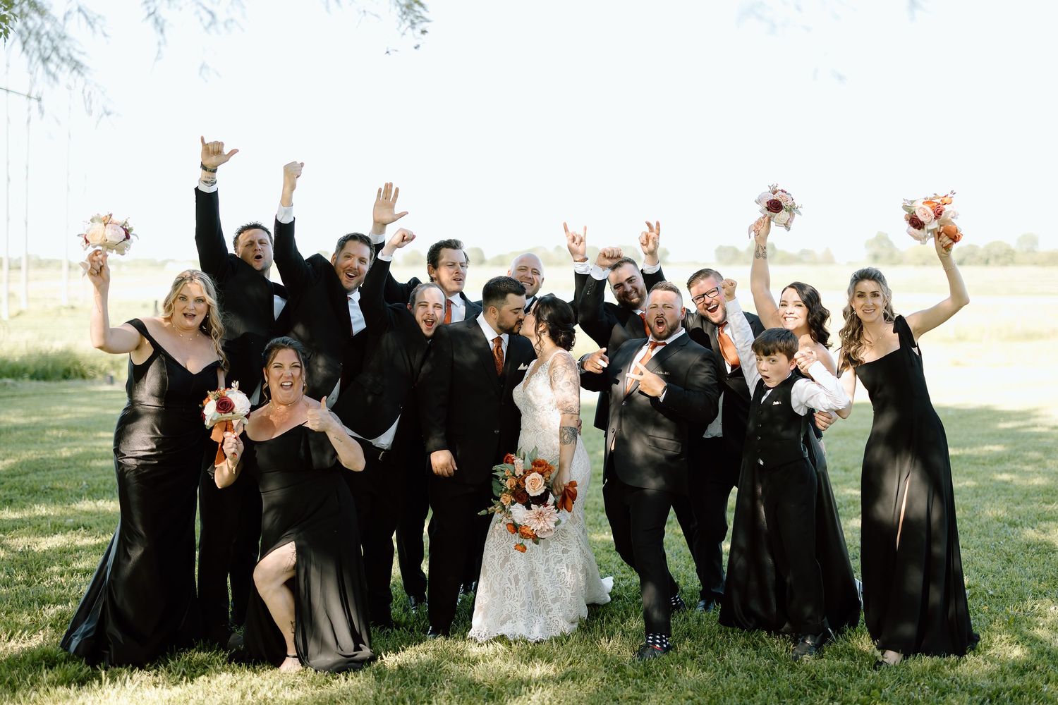 Wedding party members in black and gray attire jump joyfully for a celebratory outdoor group photo.