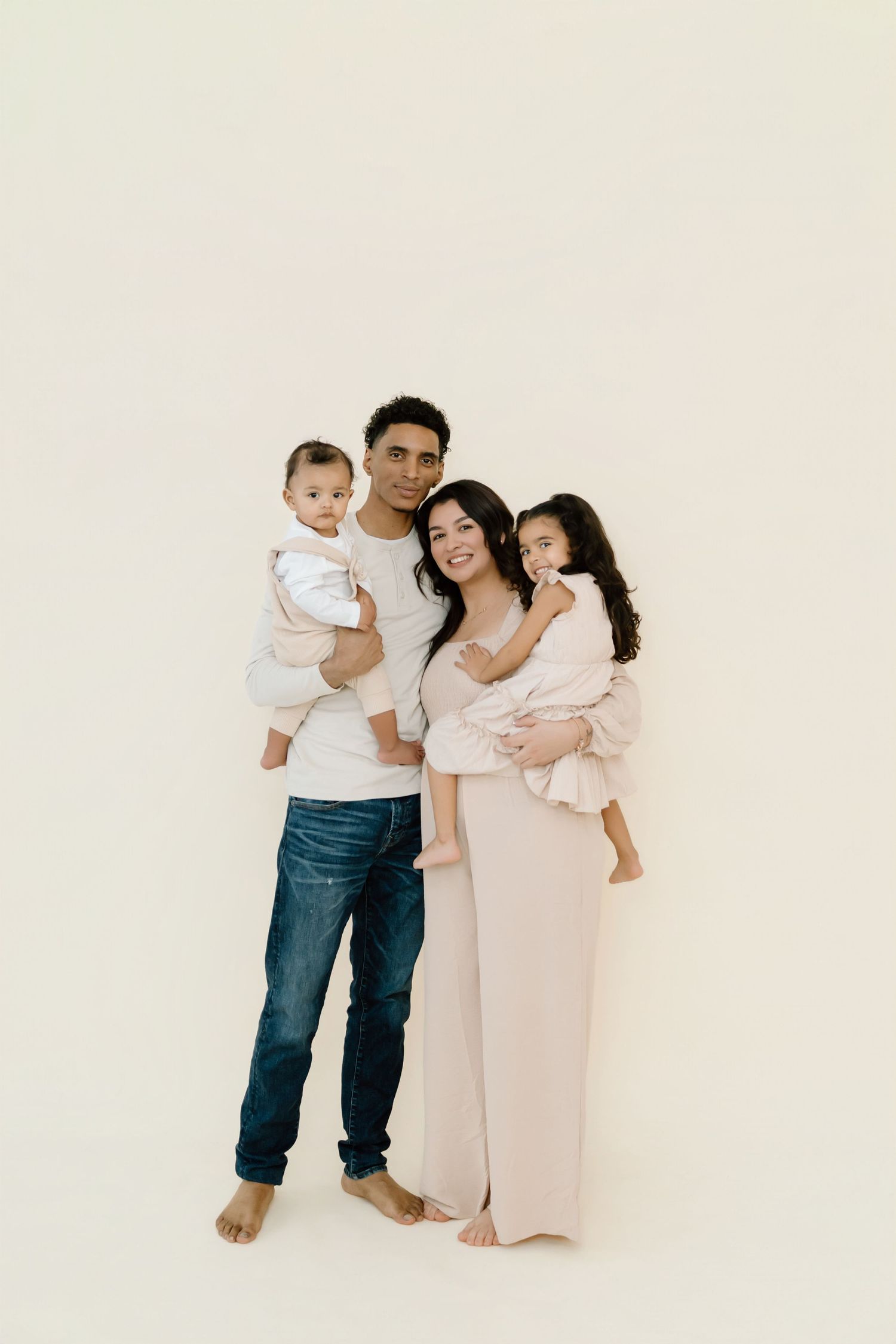 Family wearing white and beige outfits poses together against a light neutral background in a minimalist studio setting.