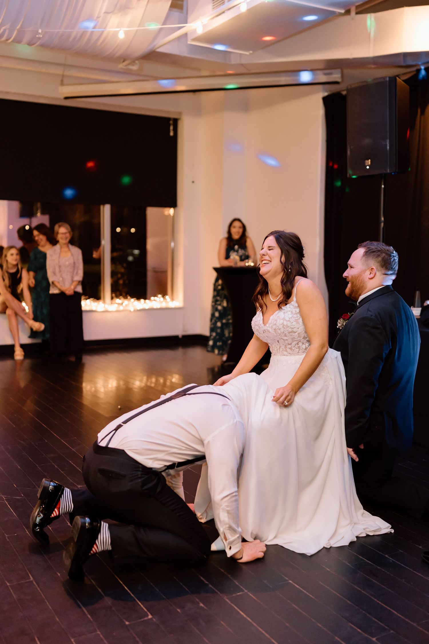 Wedding reception moment during garter toss tradition on dark hardwood dance floor with guests watching in background.