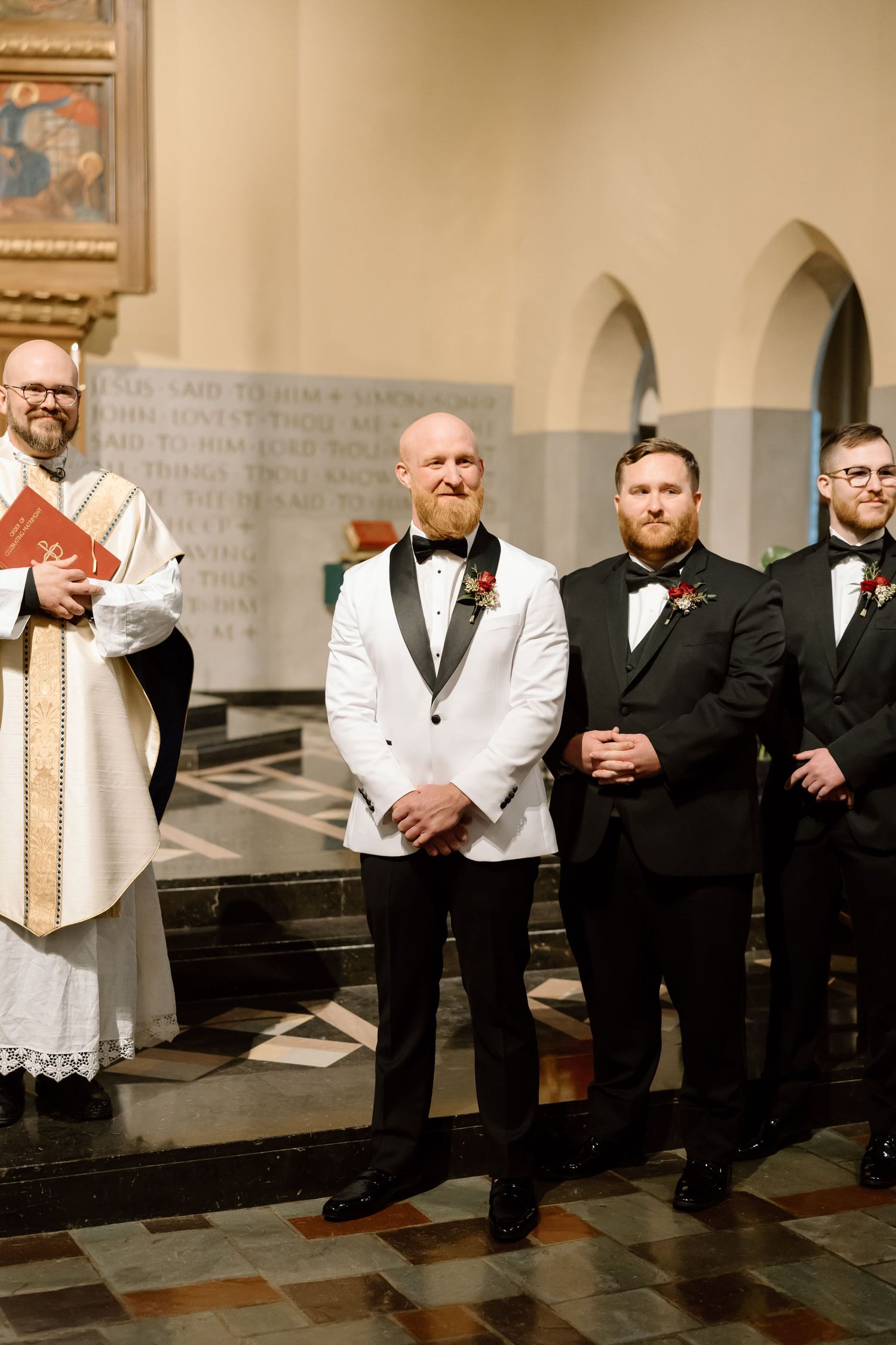 A line of clergy members in white robes and black suits stand in a church with tiled floors and stone walls.