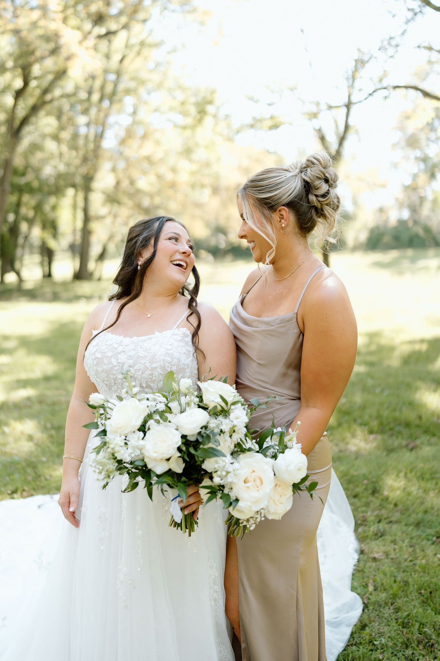 Wedding party members in white and beige dresses share joyful candid moments outdoors in a sunlit garden setting.