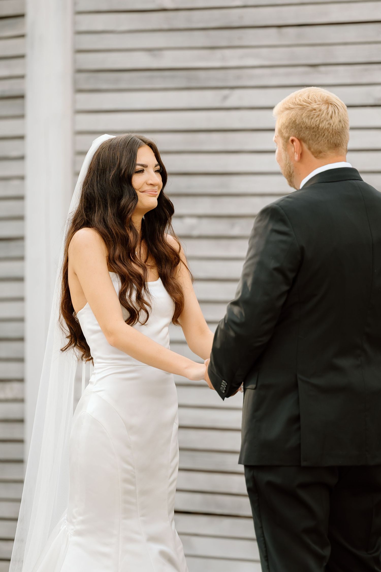 A bride in a white wedding dress and long dark hair holds hands with a groom in a black suit against a gray wall.