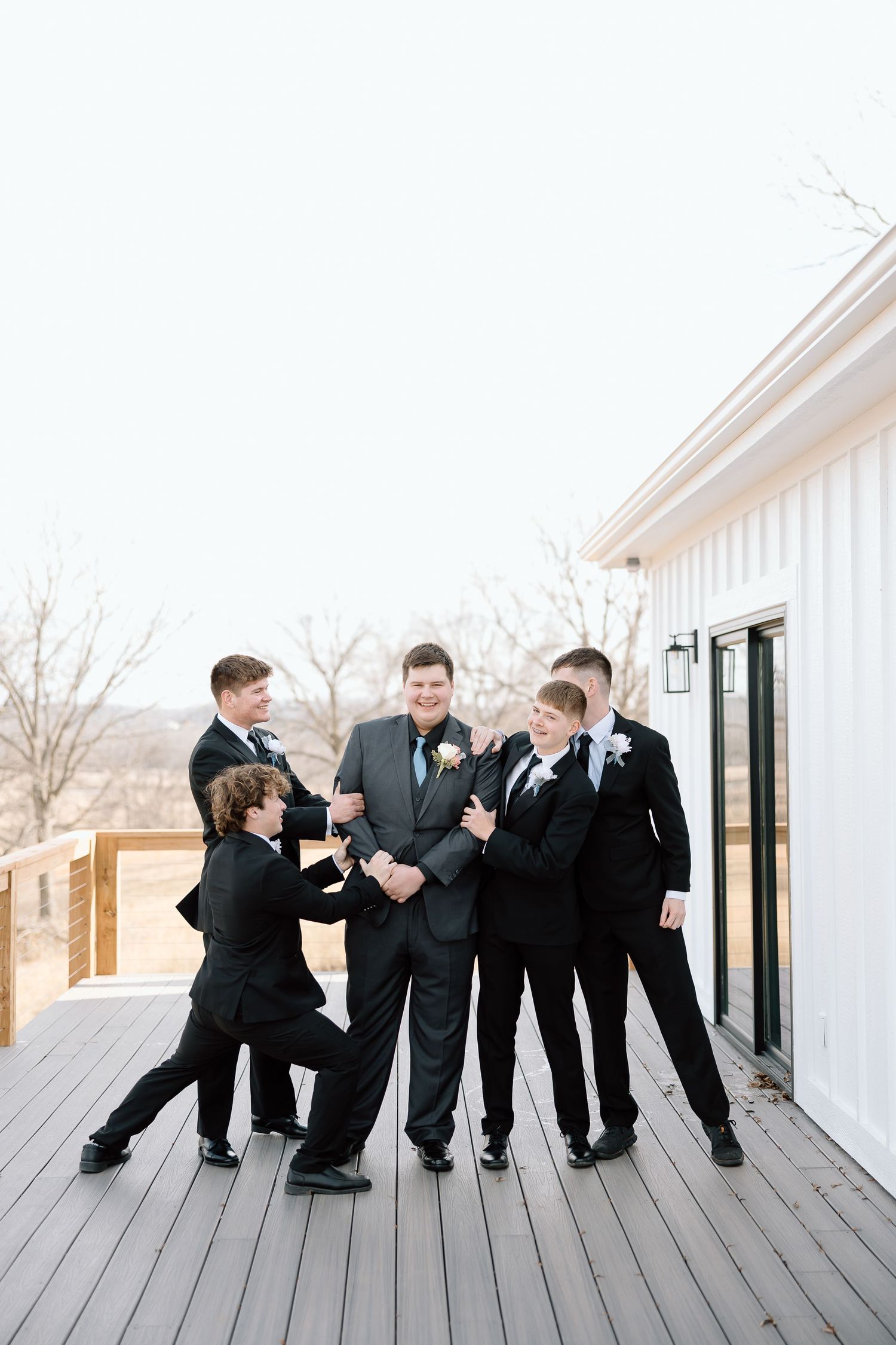 A group in formal black suits sharing a lighthearted moment on a white deck during a wedding celebration.