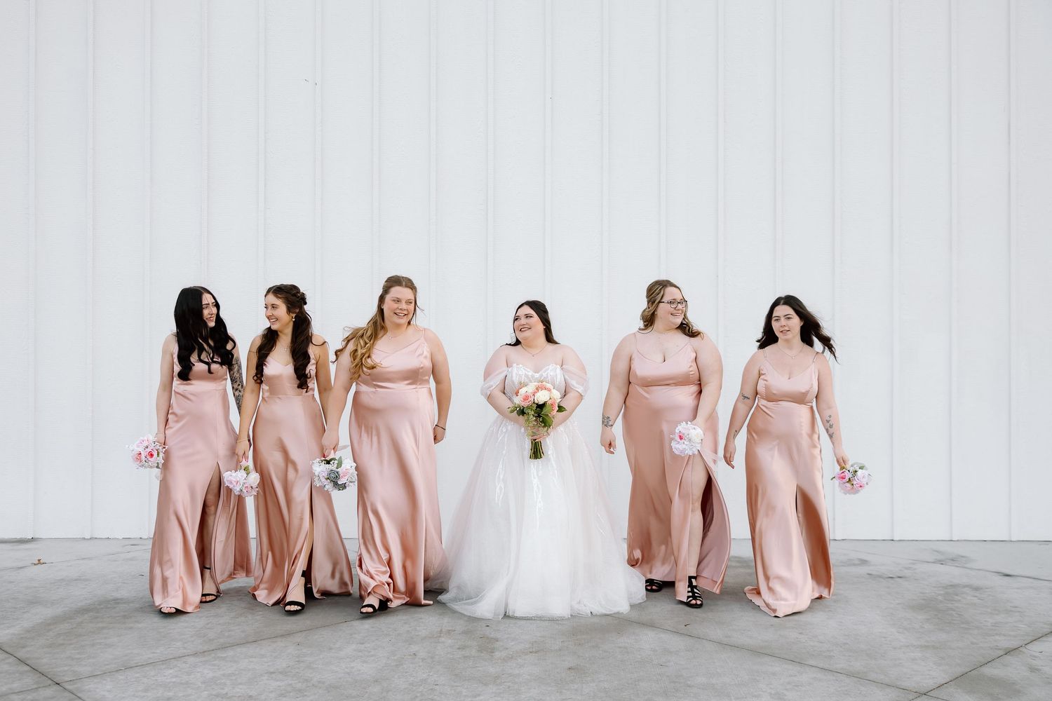 Bridal party in blush pink dresses and white wedding gown pose together in elegant indoor setting.
