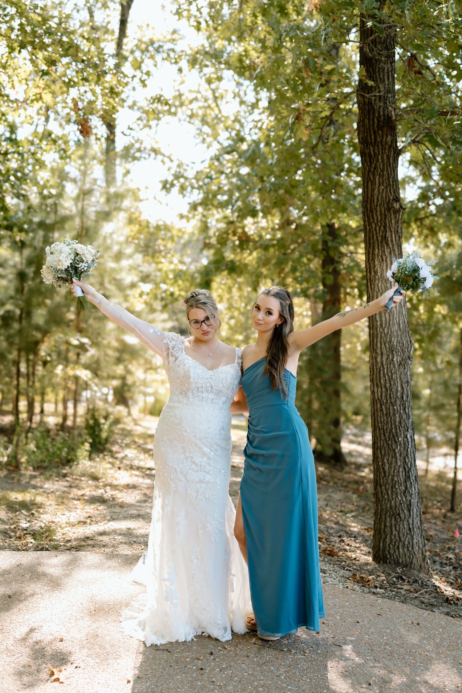 Two people in white and teal dresses dance joyfully together on a sunlit forest path surrounded by tall trees.