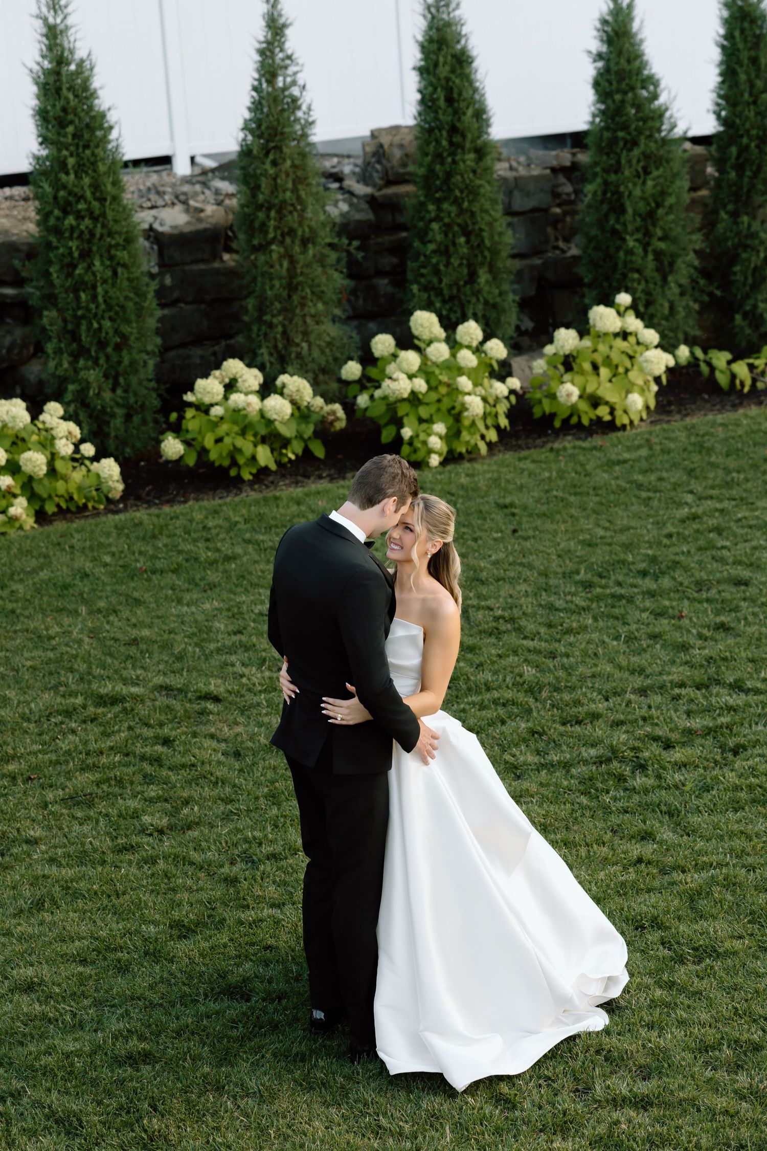 Bride and groom share a romantic kiss on lush green lawn with white hydrangeas and tall evergreen trees backdrop.
