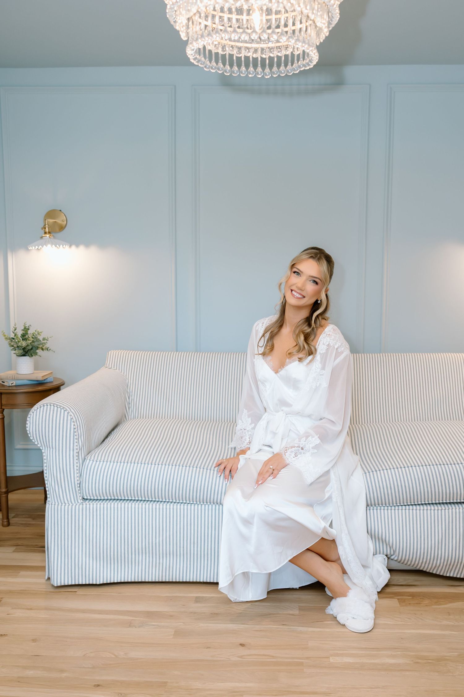 Smiling bride in white robe and sneakers sitting on striped sofa in elegant blue room with crystal chandelier.