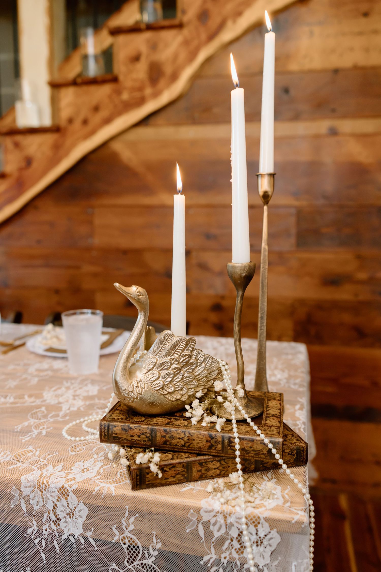 Gold swan figurine on vintage books with white taper candles and pearl beads on lace tablecloth in rustic wood interior.