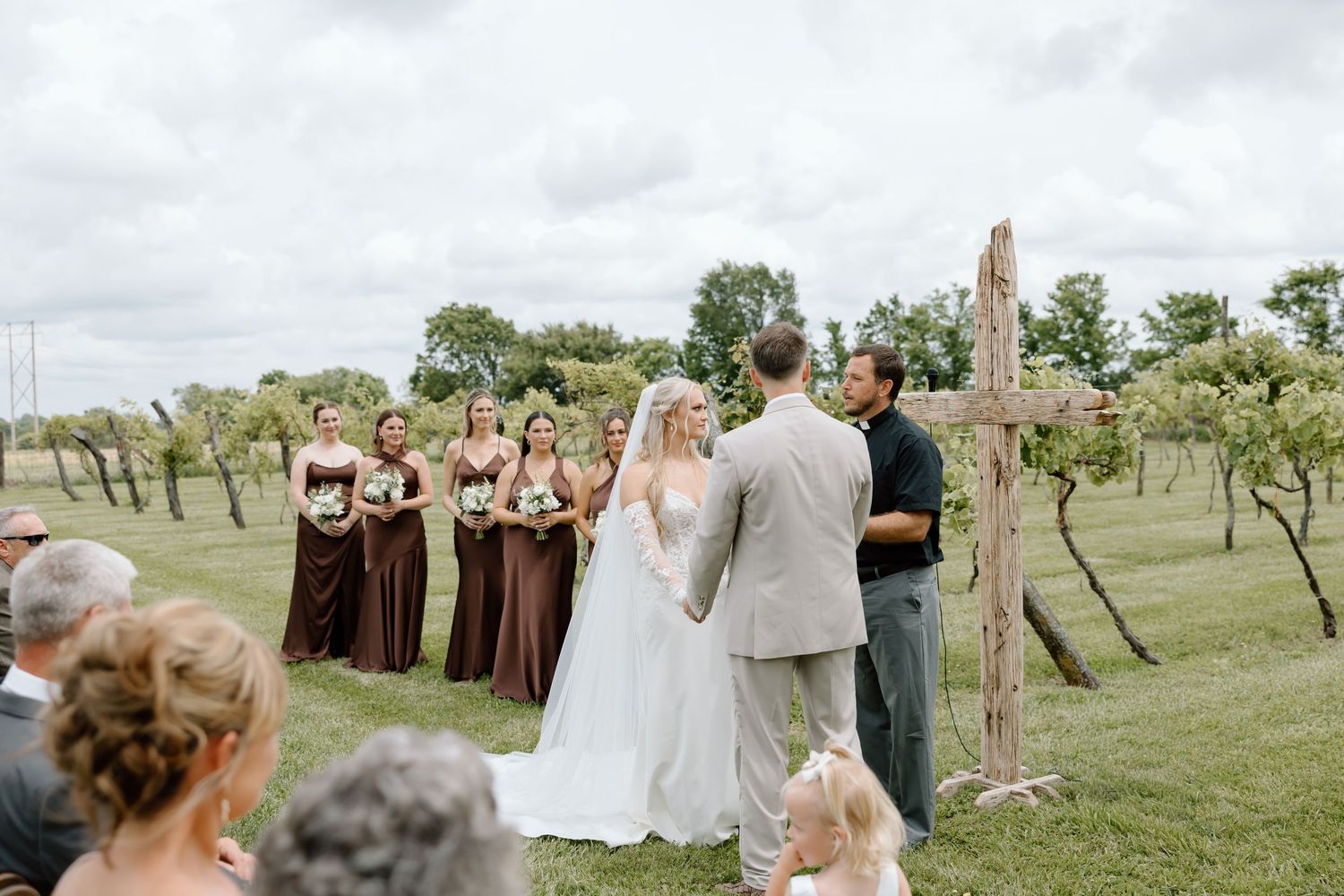 Outdoor vineyard wedding ceremony with bridesmaids in burgundy dresses, bride and groom at rustic wooden cross altar.