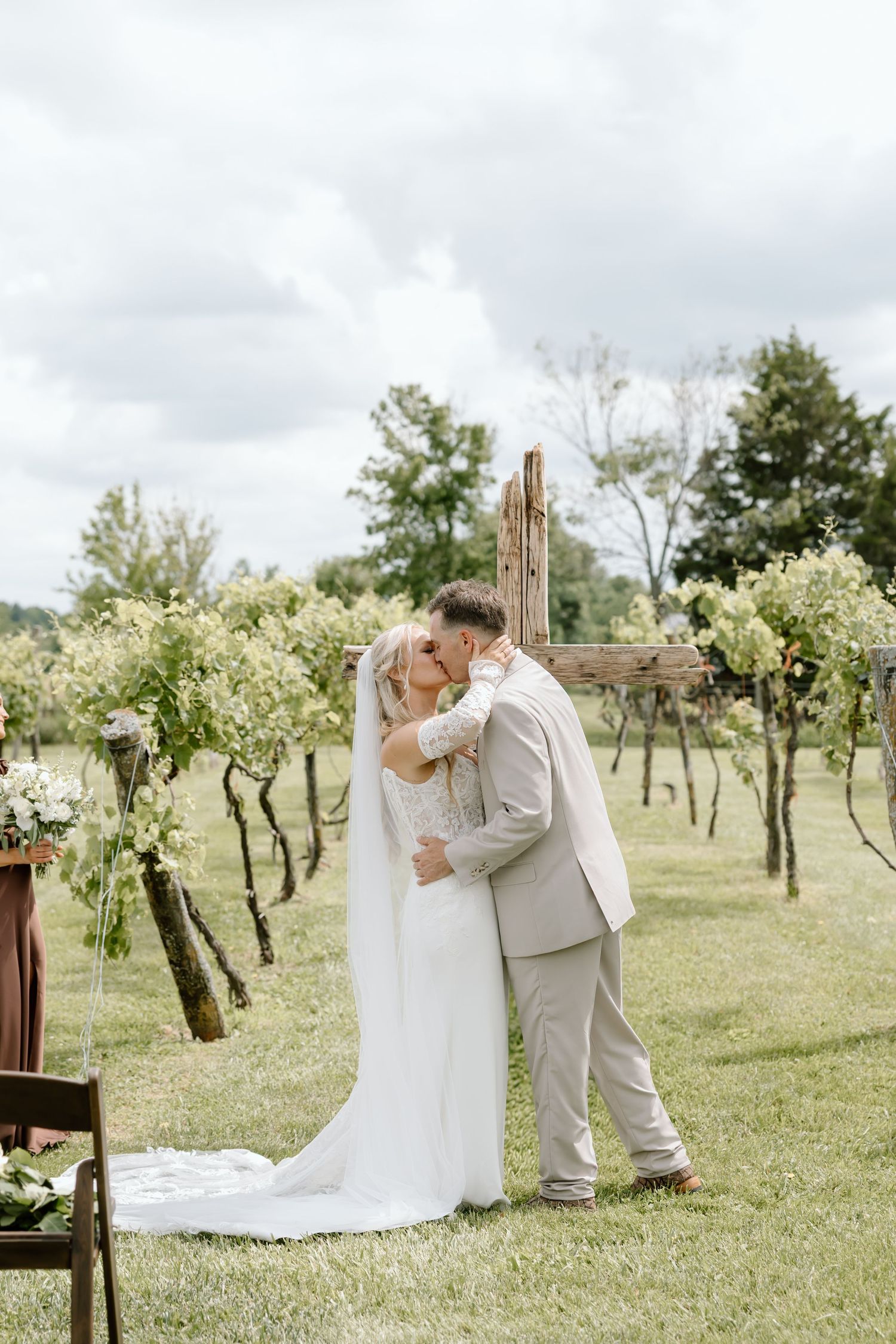 Bride and groom share first kiss at outdoor vineyard wedding ceremony with rustic wooden cross and floral greenery backdrop.