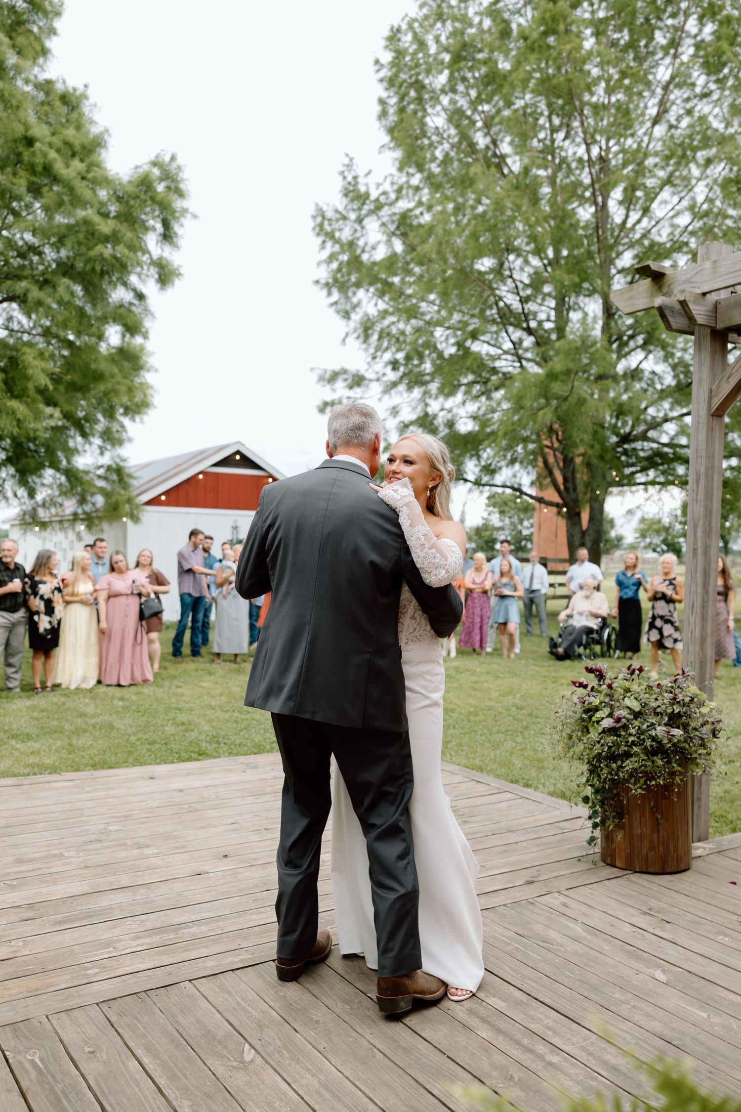 Bride and father share first dance on outdoor wooden deck at rustic farm wedding with guests watching.
