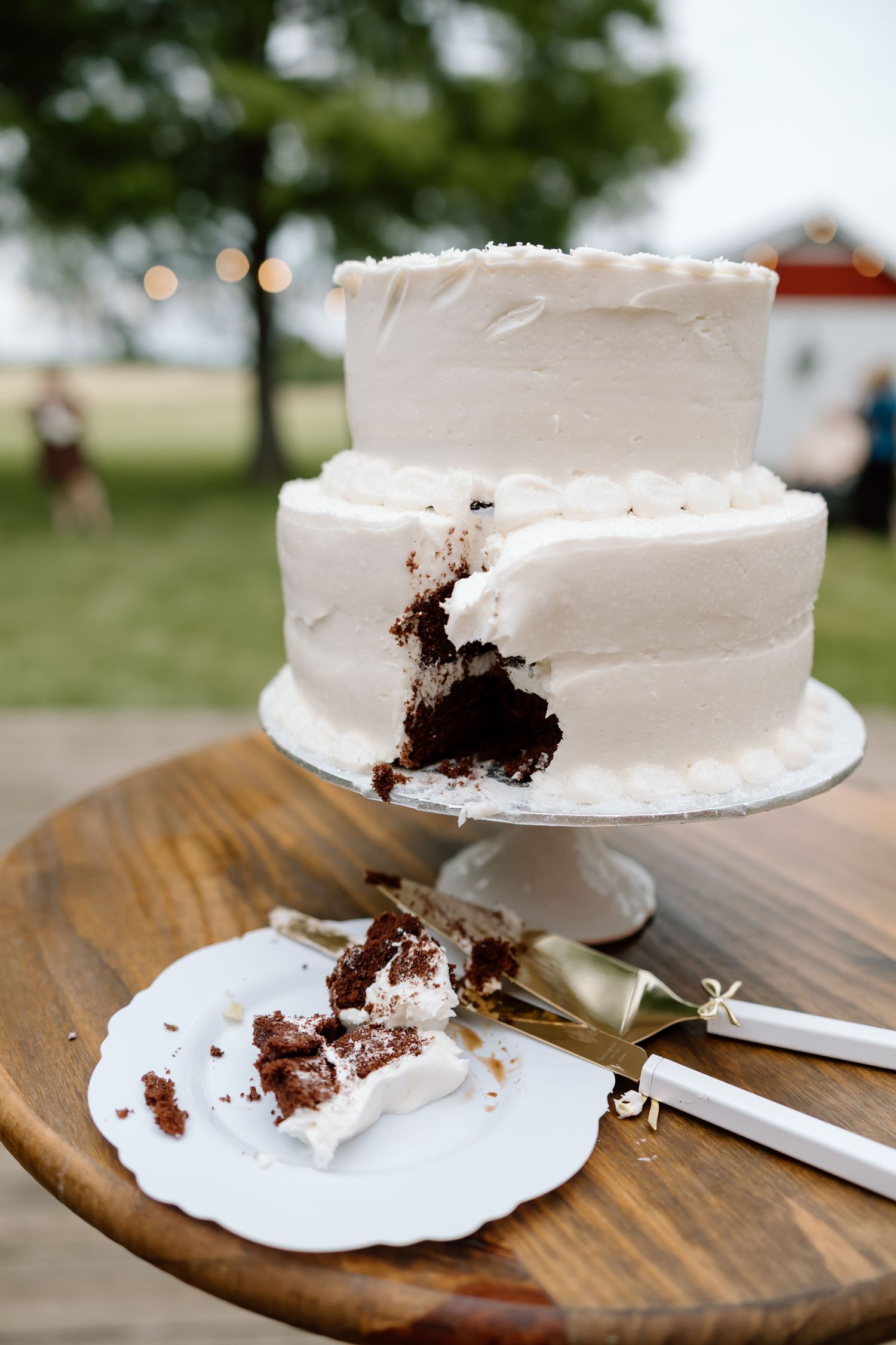 Two-tier white frosted wedding cake with chocolate interior on wooden table, with a slice served on white plate beside cake knife.