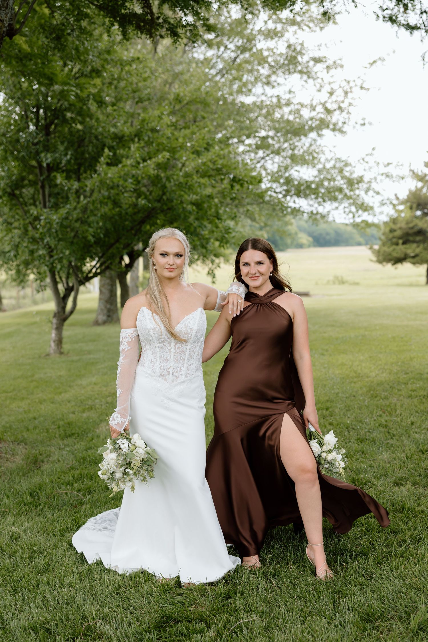 Bride in white lace gown and bridesmaid in brown satin dress holding bouquets outdoors on green lawn.