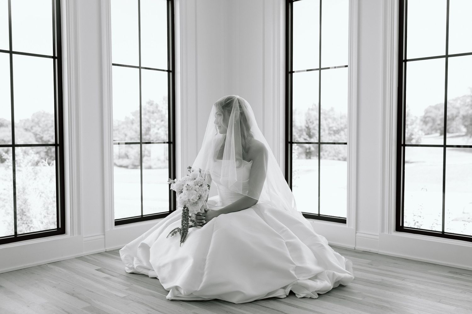 Bride in white gown and veil sits on floor by large windows holding bouquet, black and white photo.