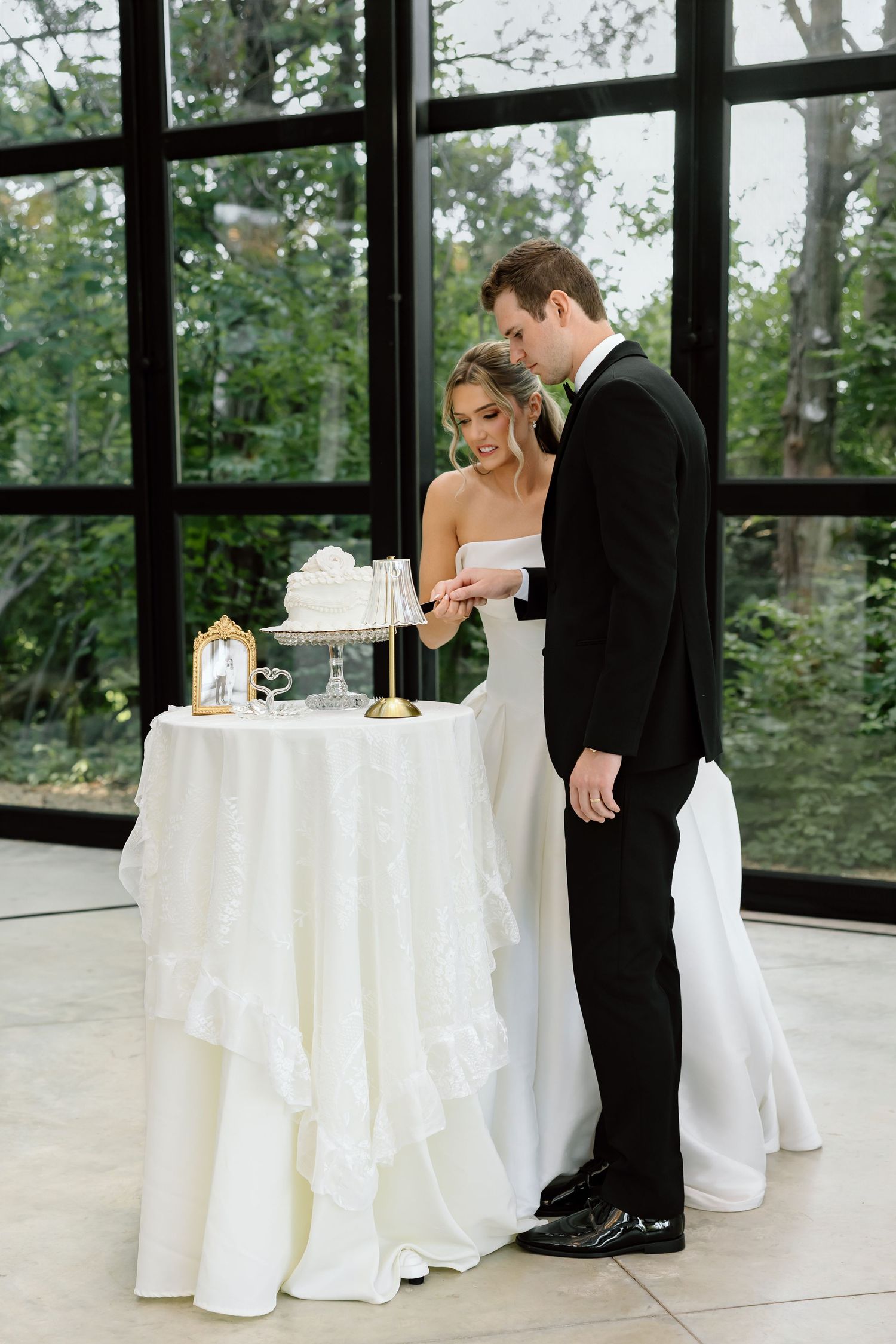 Bride and groom cutting their white wedding cake together at an elegant indoor venue with large windows.
