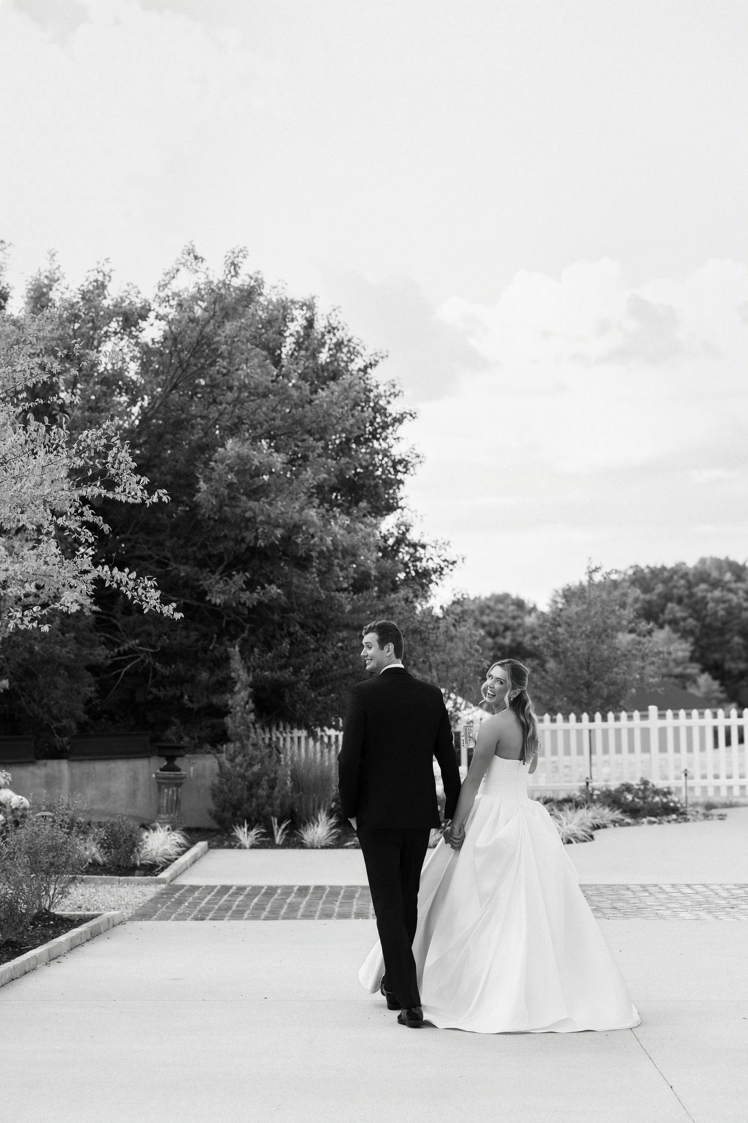 Black and white photo of a bride and groom walking hand in hand outdoors, with lush trees and a white fence backdrop.