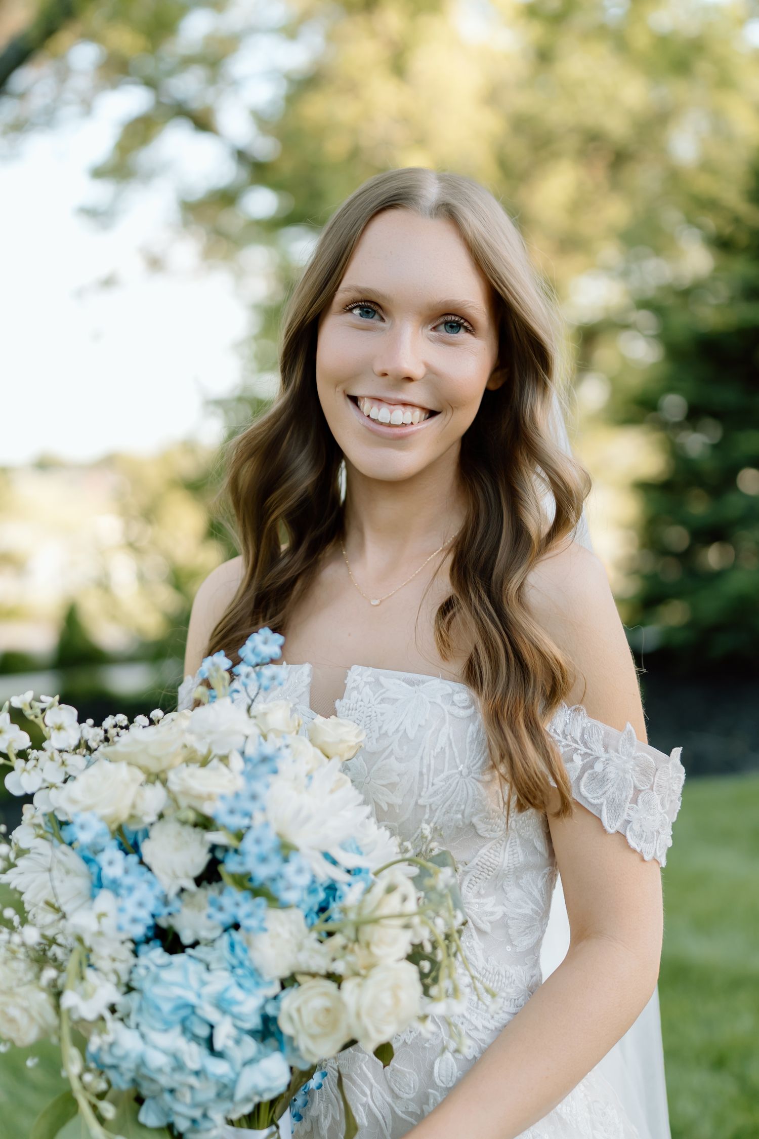 Smiling bride in off-shoulder lace gown holding blue and white floral bouquet outdoors.