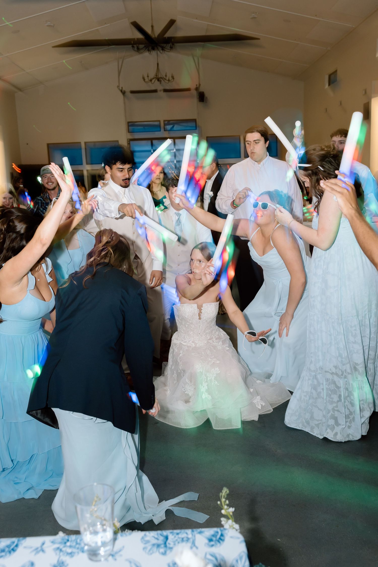 Bride surrounded by wedding guests waving colorful LED glow sticks on the dance floor during reception.