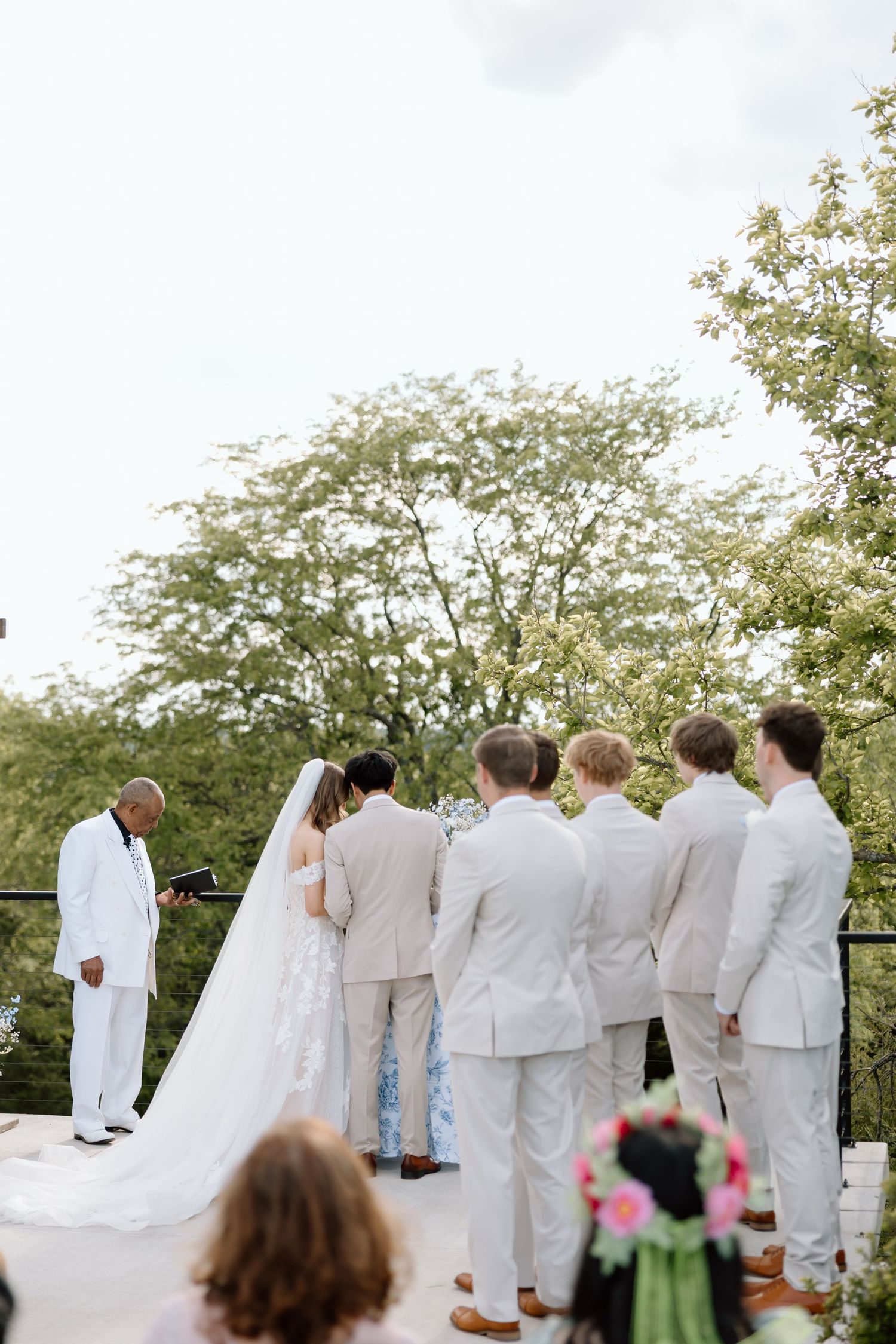 Outdoor wedding ceremony on a terrace with bride, groom, groomsmen in tan suits, and officiant in white suit, surrounded by lush trees.