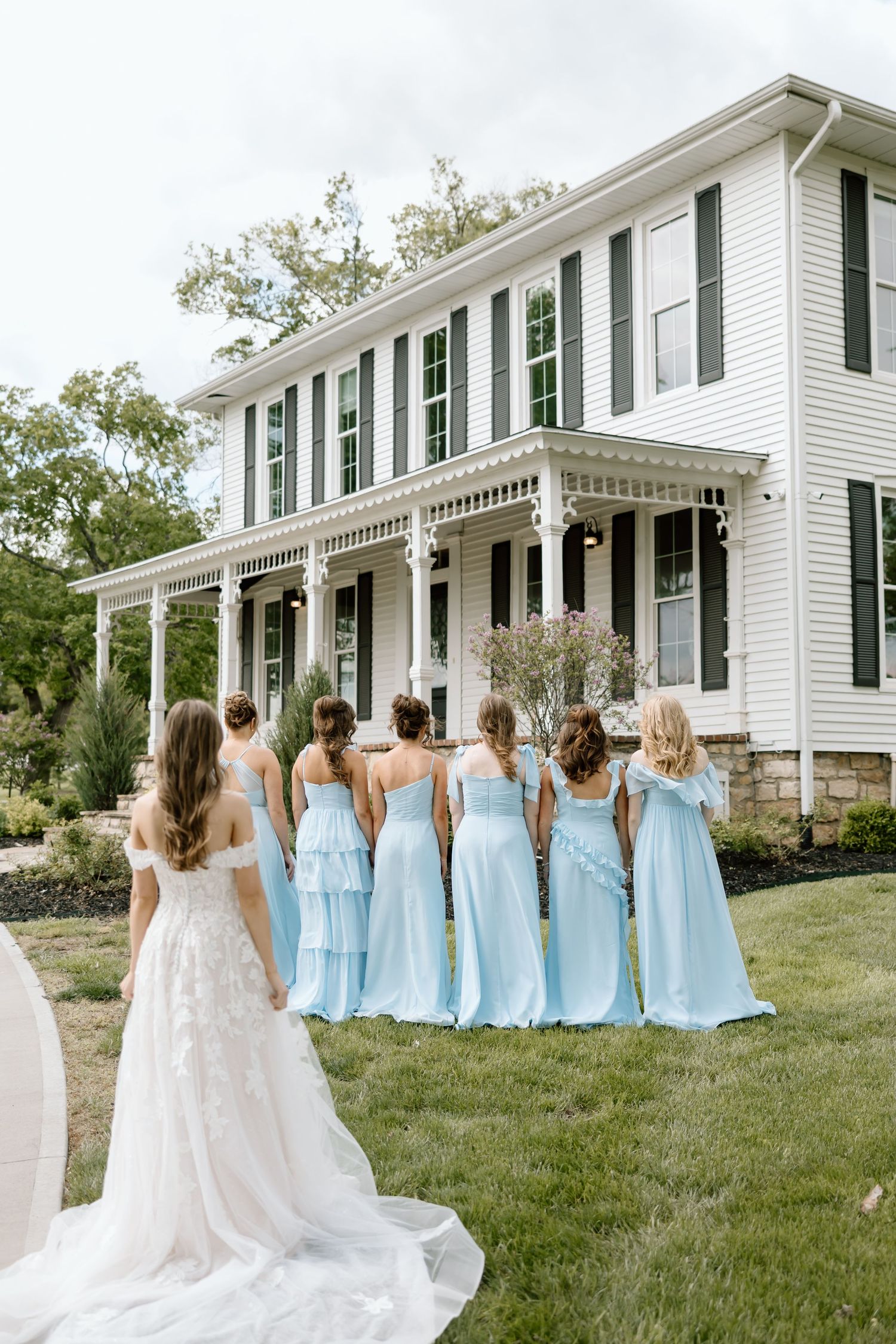 Bride in white lace gown approaches bridesmaids in light blue dresses standing before a white colonial wedding venue.