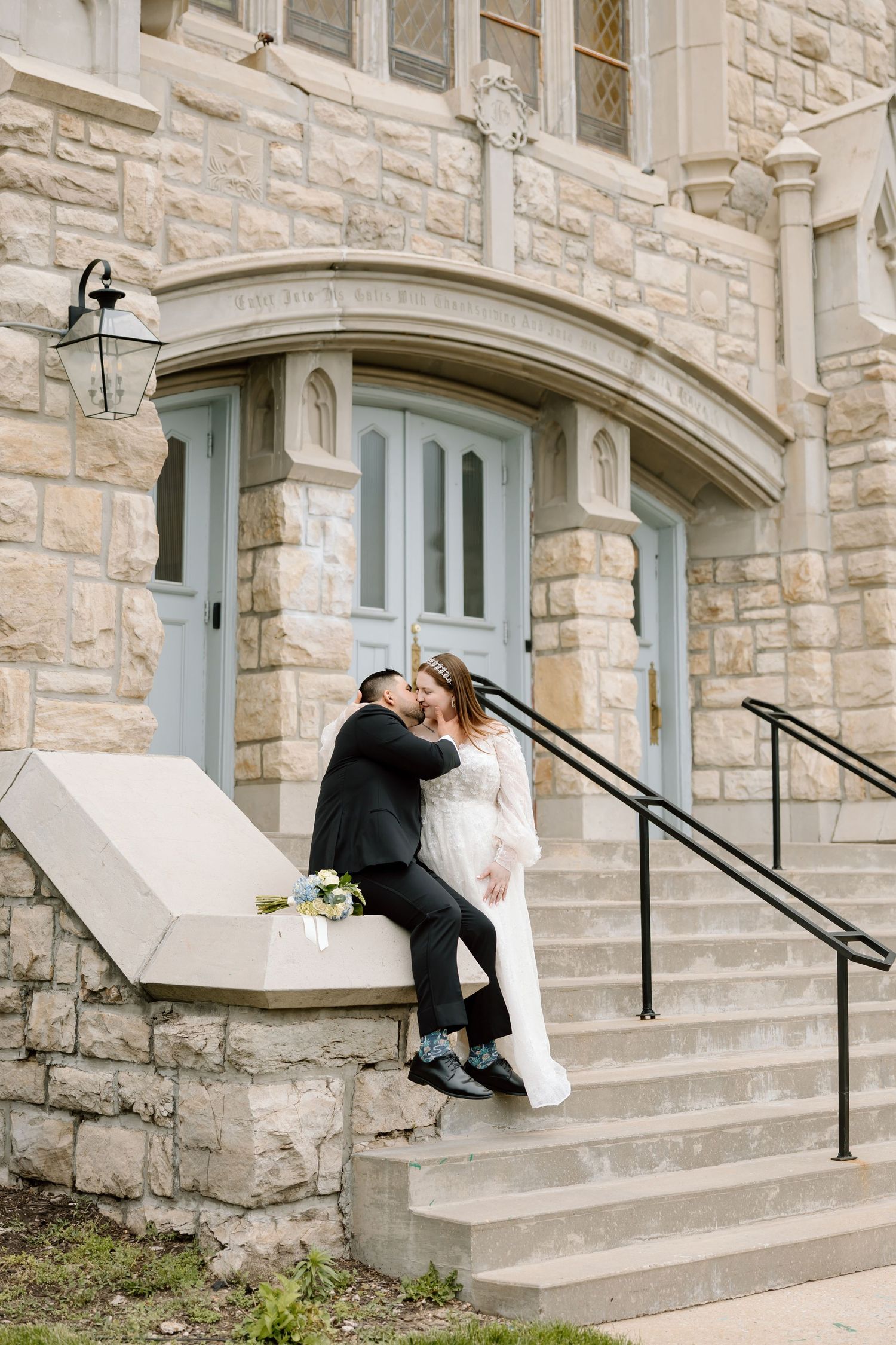 Bride and groom share a kiss on stone steps outside a historic building on their wedding day.