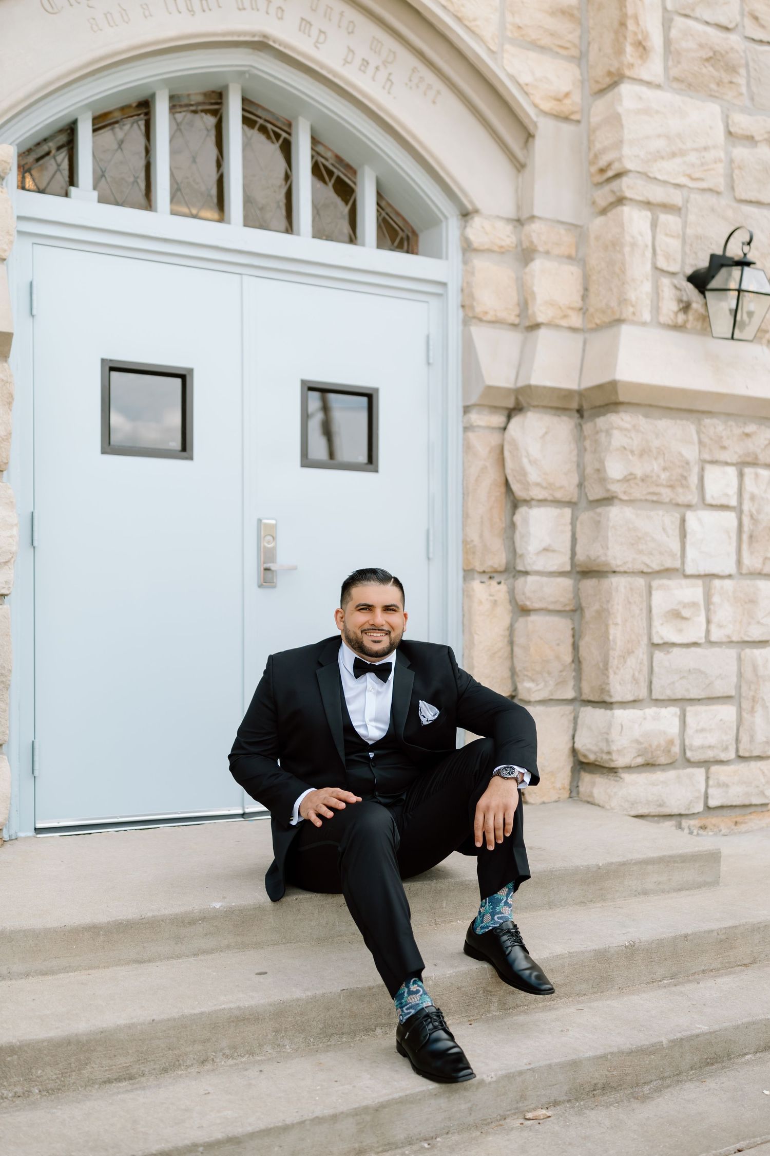 Groom in black tuxedo sitting on church steps smiling, wearing patterned socks and black dress shoes.