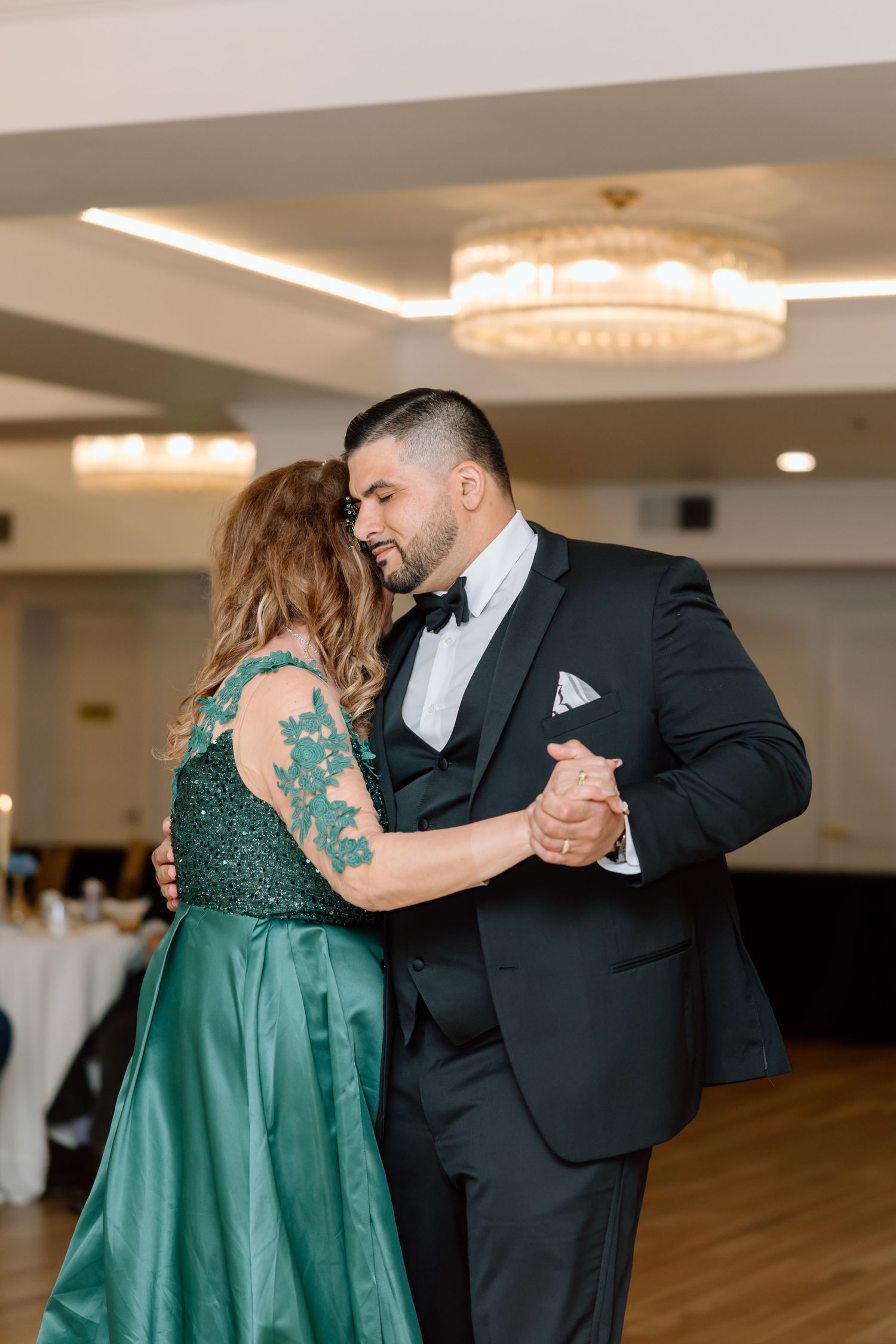 A groom in a black tuxedo shares a tender dance with a woman in an emerald green gown at a wedding reception.