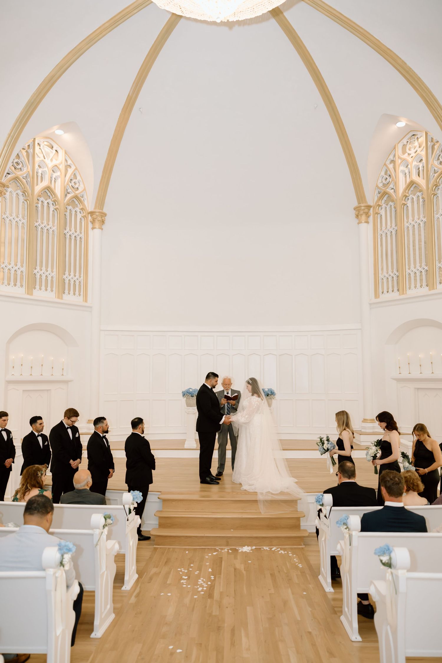 Elegant white chapel wedding ceremony with couple exchanging vows beneath a grand arched ceiling with Gothic windows.