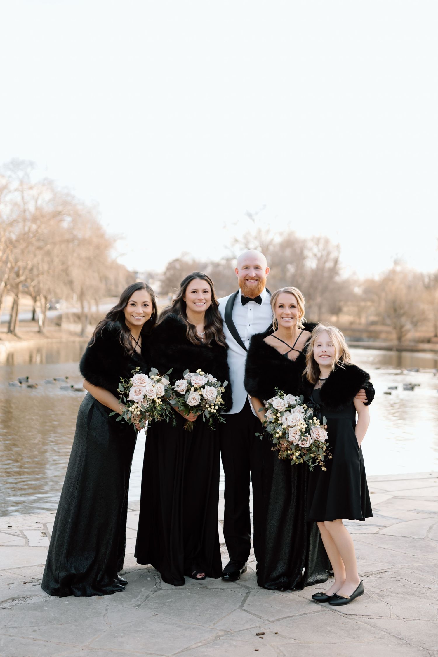 Wedding party in formal black attire poses with white floral bouquets beside a serene lake.