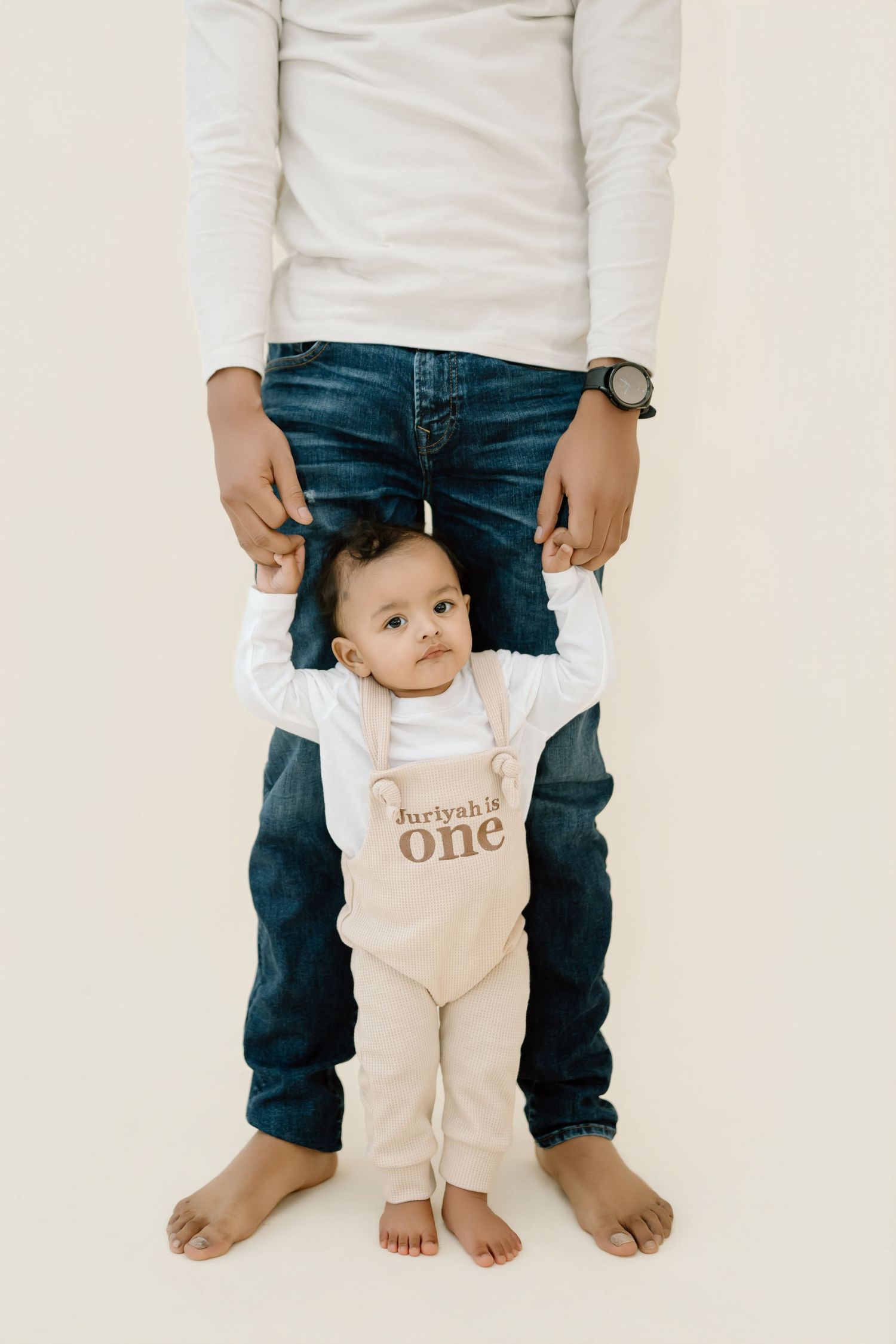 A baby in a white onesie stands supported between adult legs wearing blue jeans against a light background.