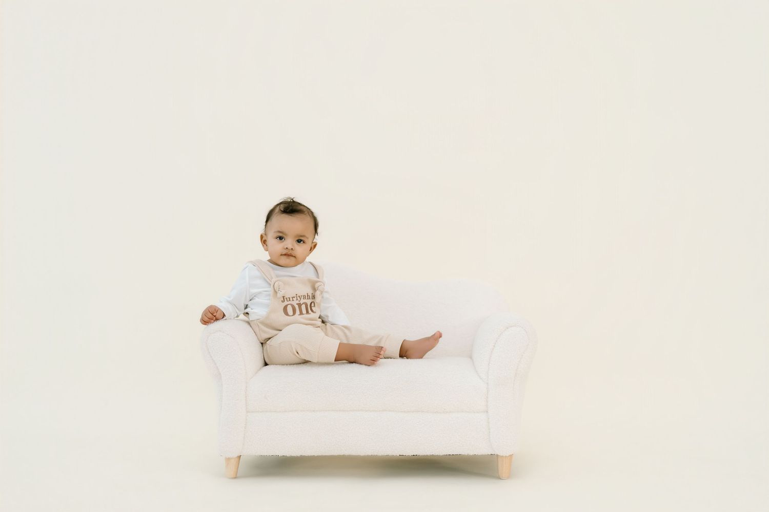 A baby in white clothing lies on white bedding during a minimalist photo session.