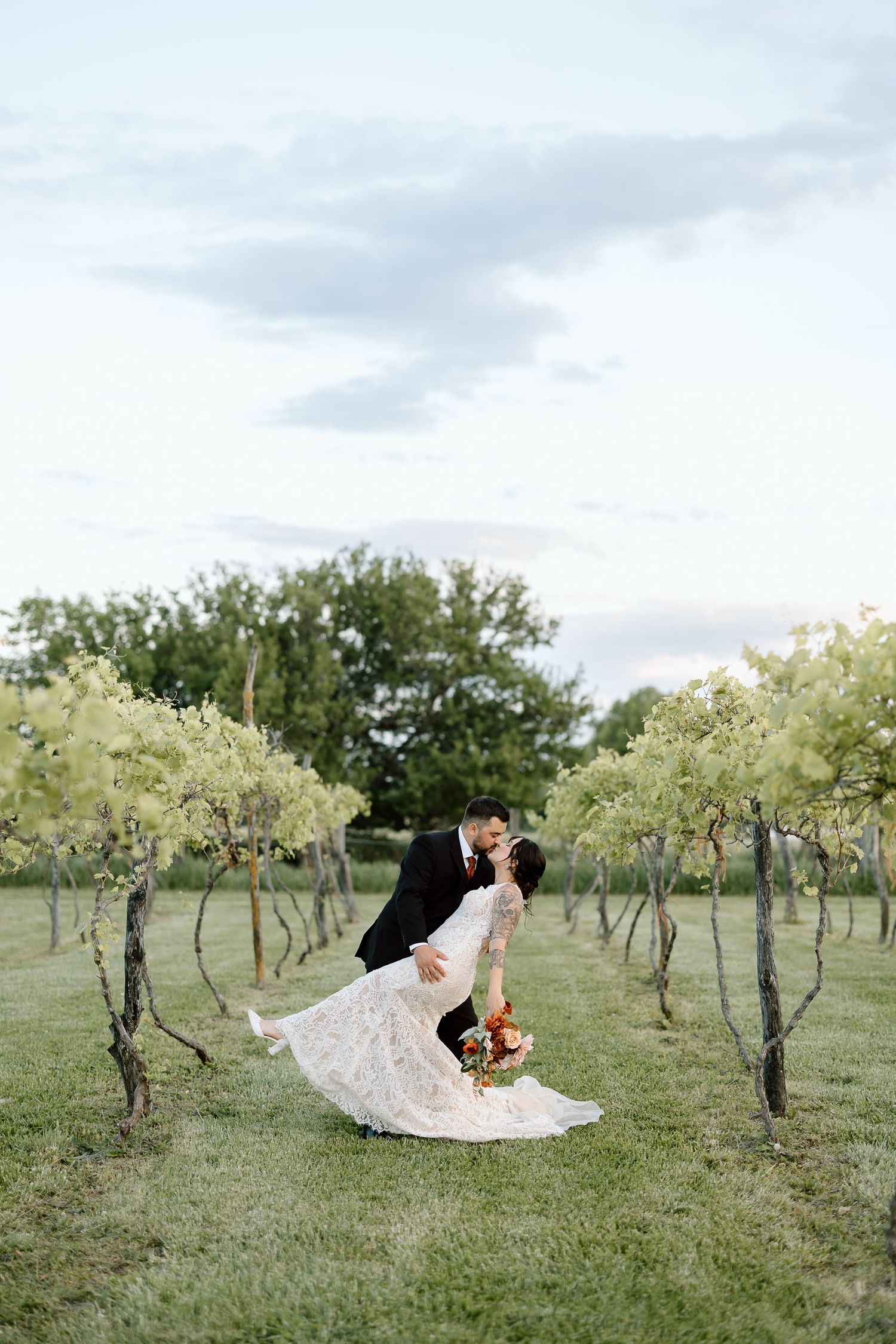 Romantic wedding couple share an intimate dip kiss in a scenic vineyard at sunset.