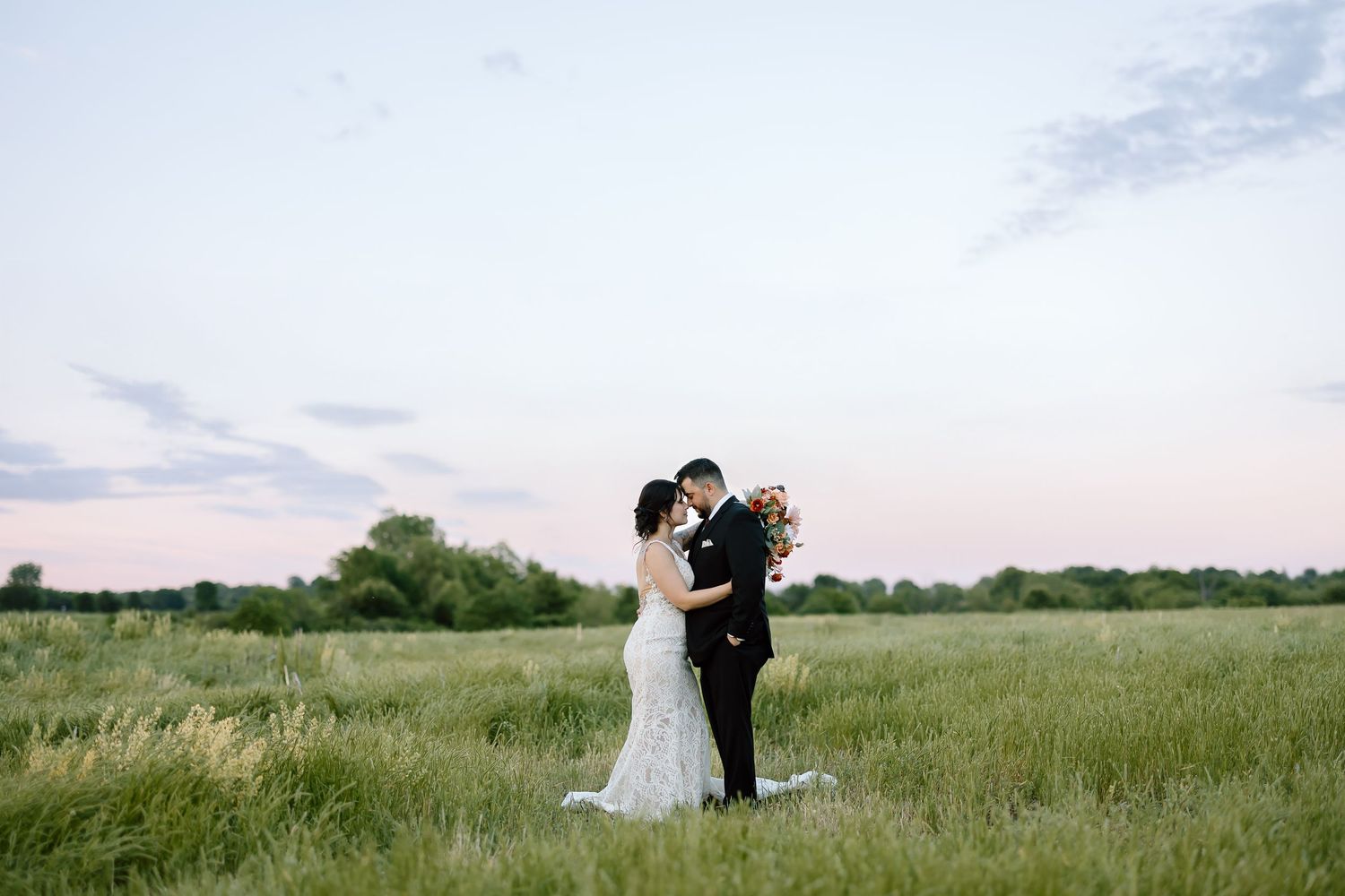 A couple stands together in an open meadow during sunset as their flowing outfits billow in the summer breeze.