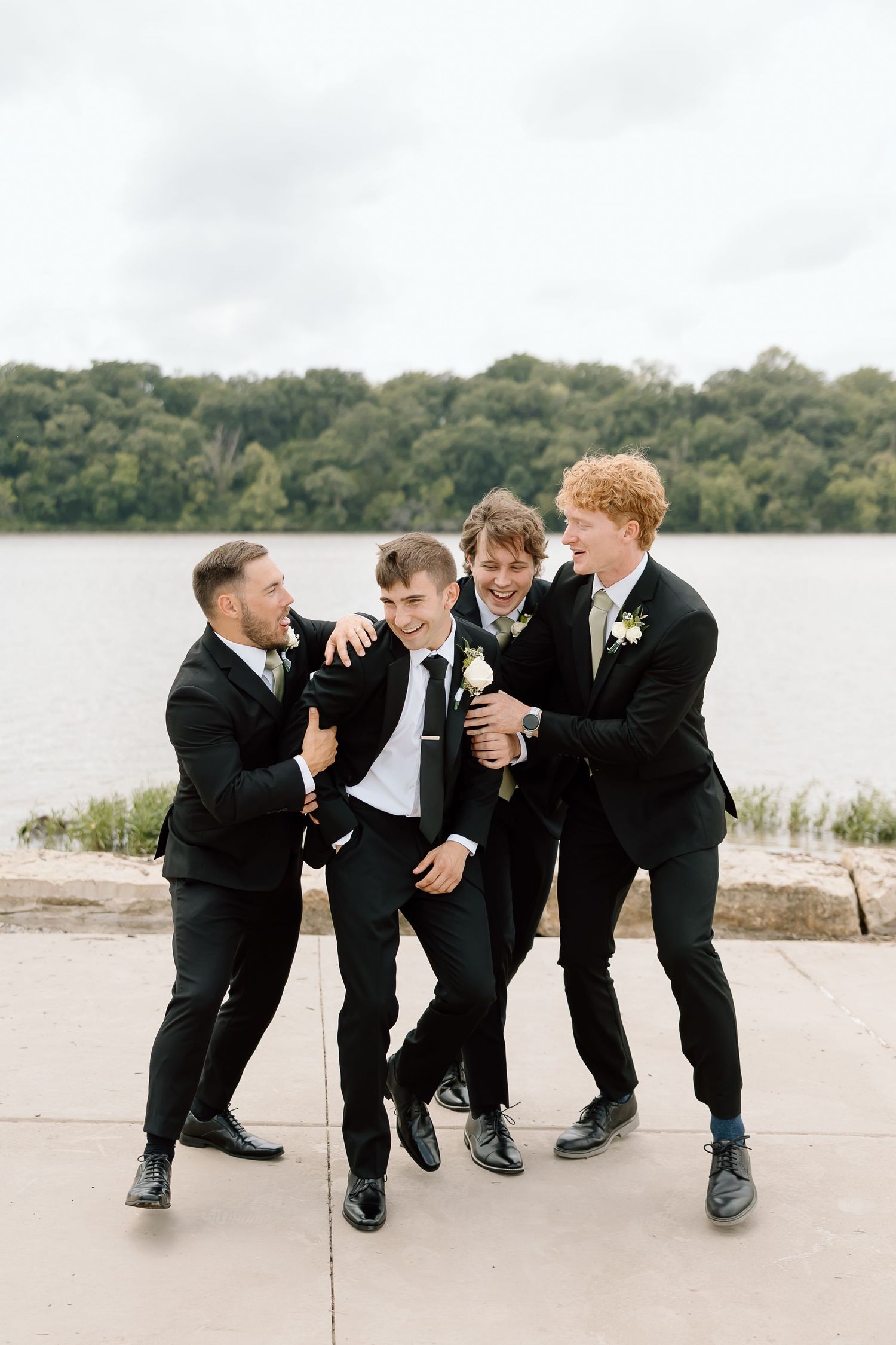 Four groomsmen in black suits laughing and joking together by a lake during wedding photos.