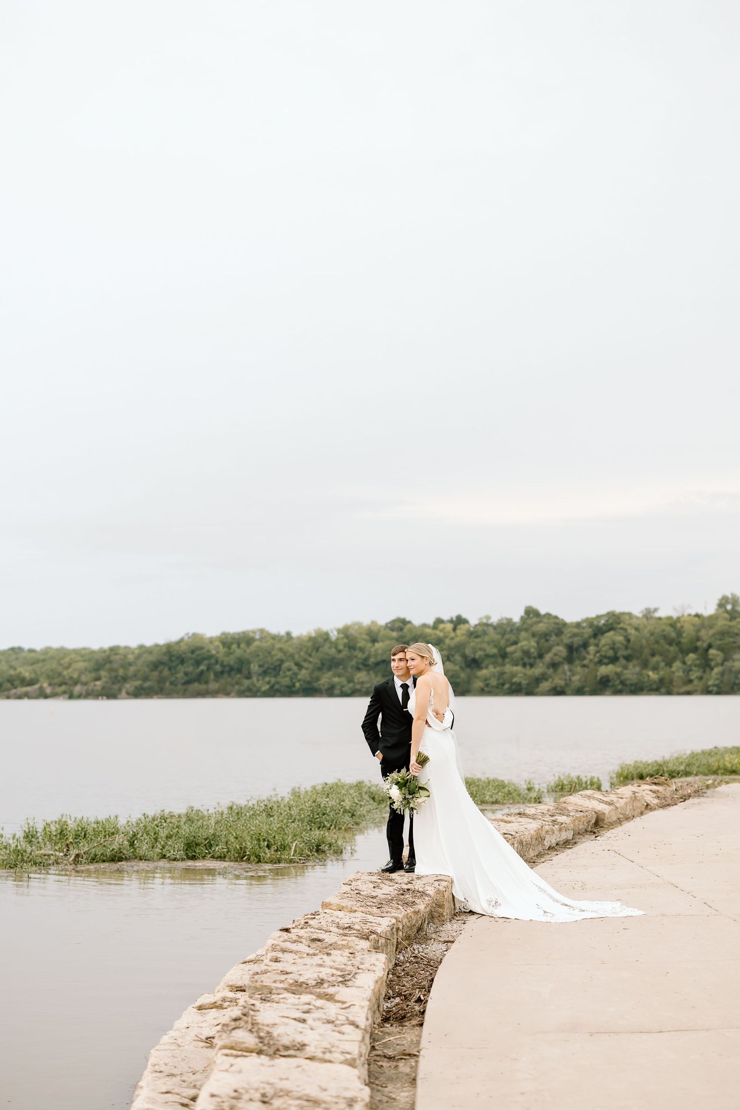 A newlywed couple embraces on a scenic pathway overlooking a serene waterfront during their wedding photoshoot.