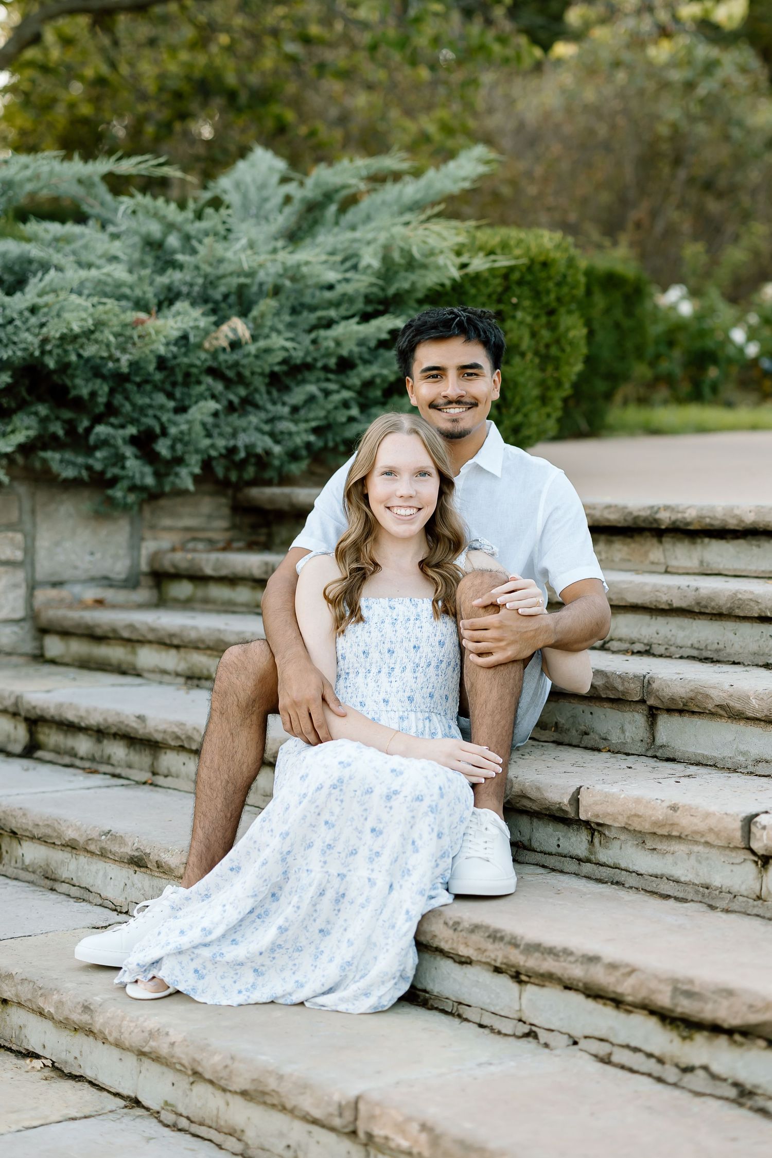 A couple poses playfully on outdoor stone steps surrounded by greenery, with the woman wearing a white floral dress.