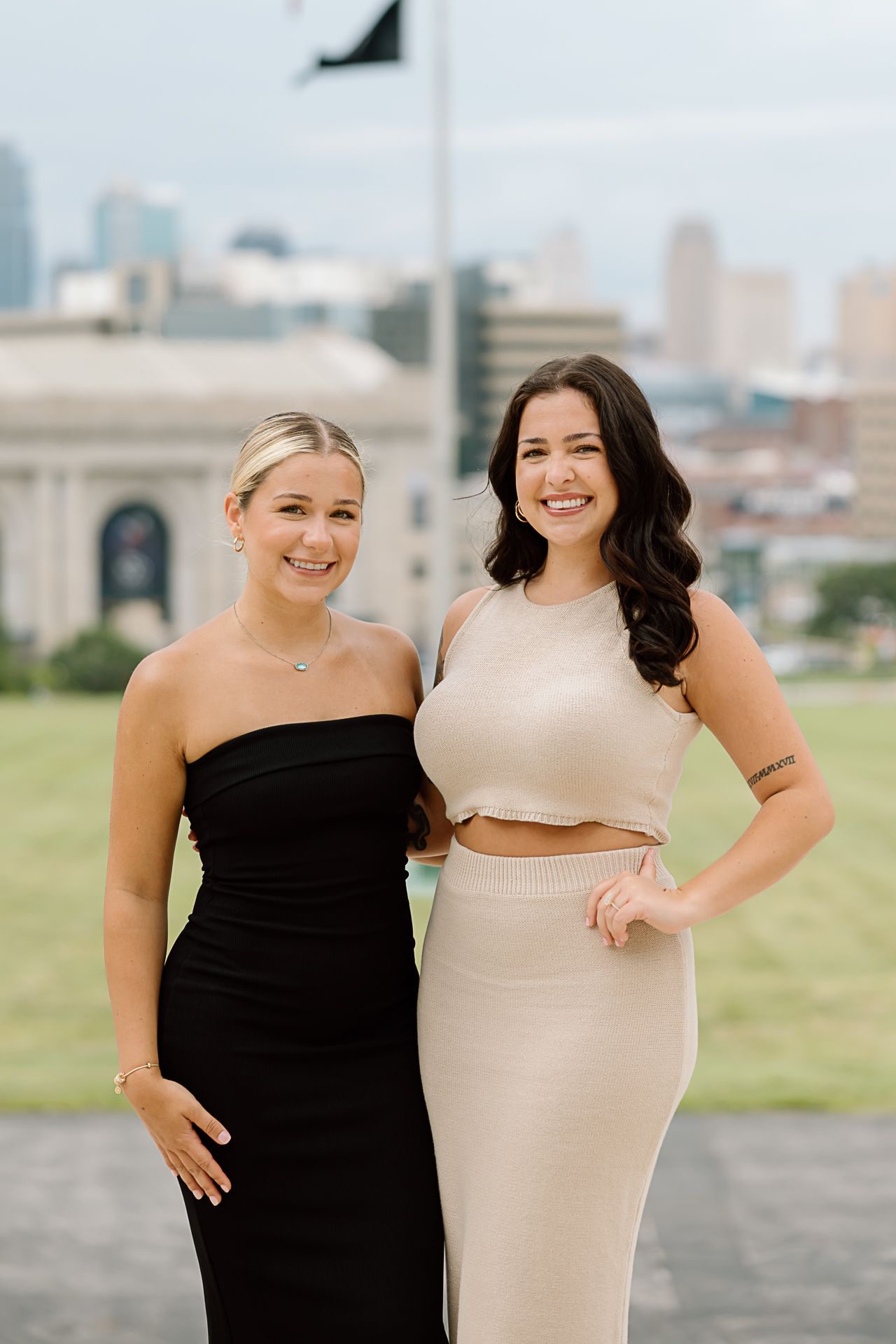 Two friends in contrasting black and beige dresses pose together outdoors with a city skyline in the background.