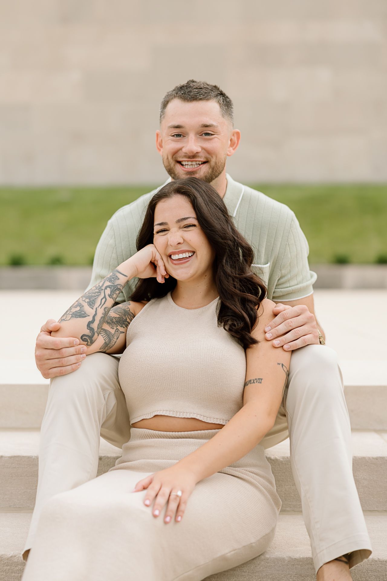 A couple laughs together while relaxing on outdoor steps in matching cream-colored outfits.