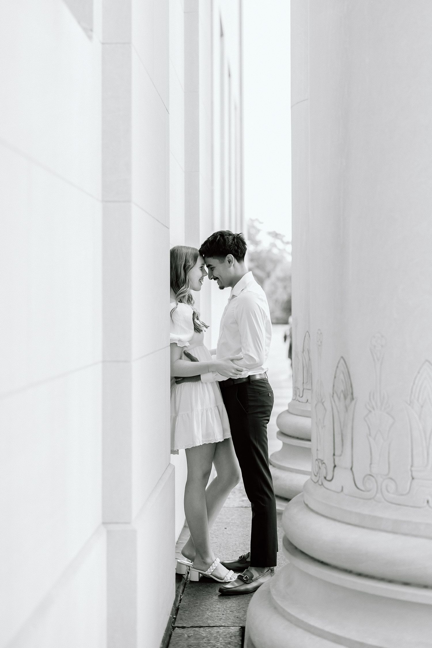 A romantic black and white photo of a couple embracing between elegant white columns in classic architectural setting.