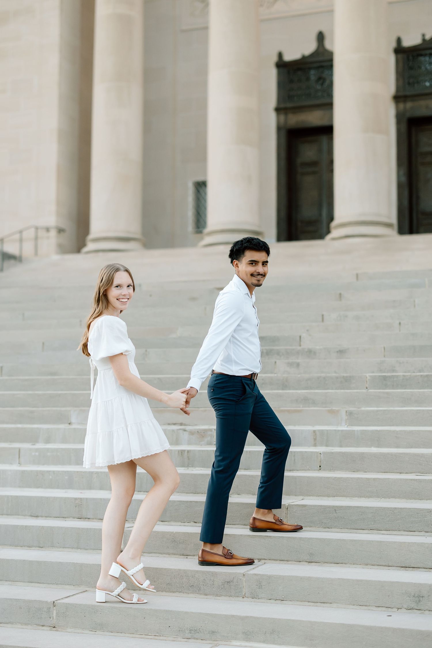 A couple playfully runs up white marble steps in formal attire during a photo session.