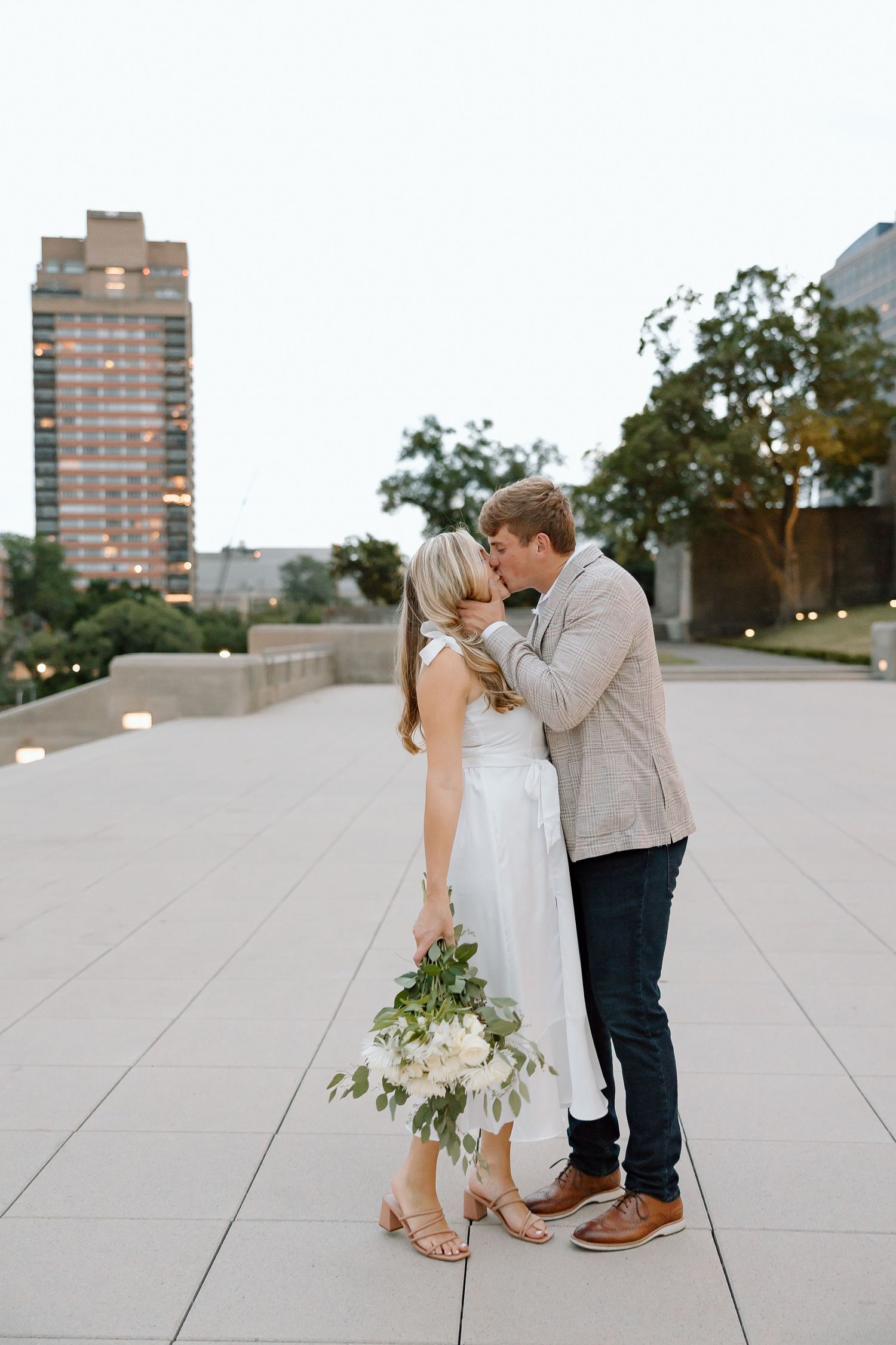 A romantic couple shares a tender moment on a modern concrete plaza with urban buildings in the background.