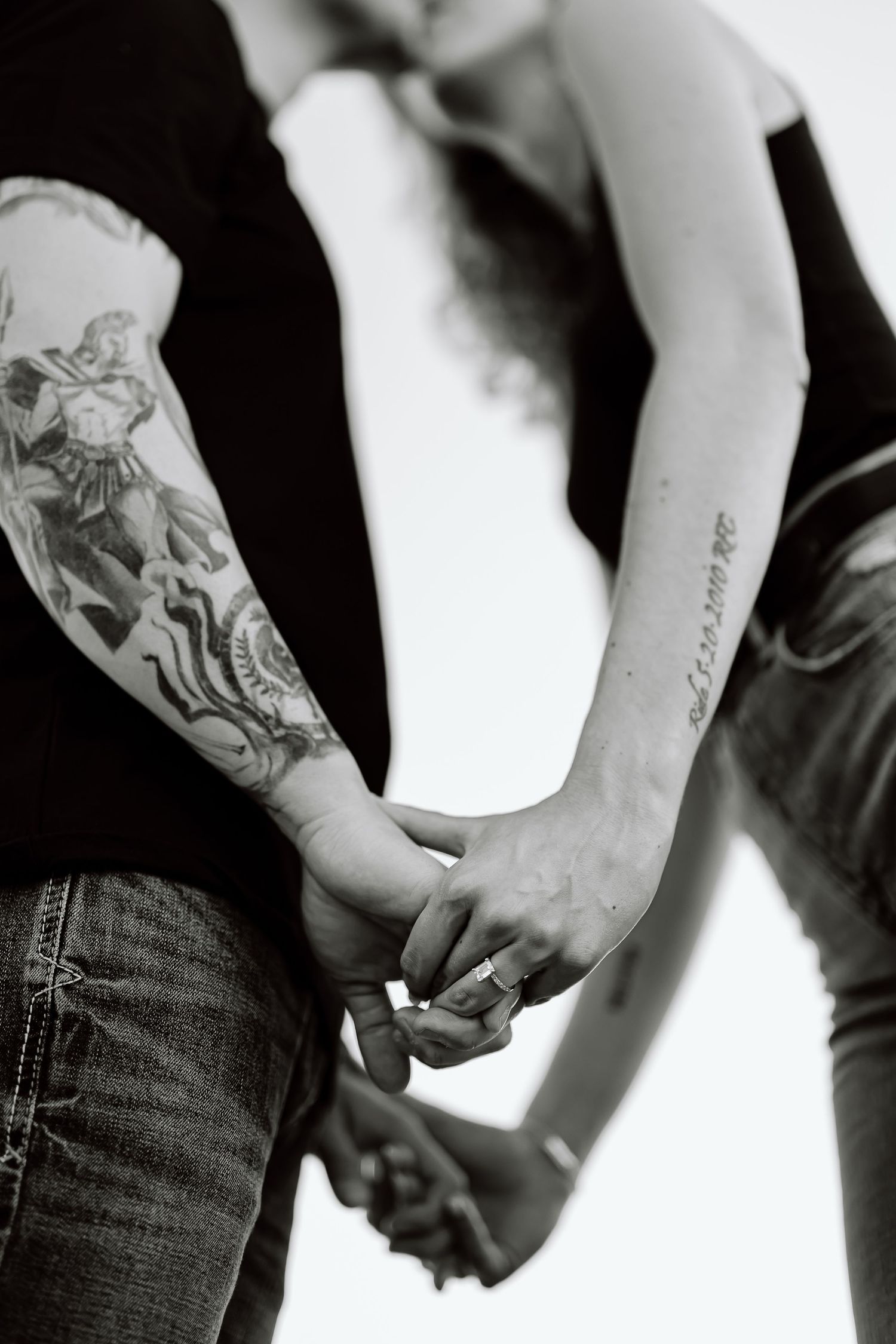 Black and white image of two people holding hands, with one arm featuring detailed sleeve tattoos.