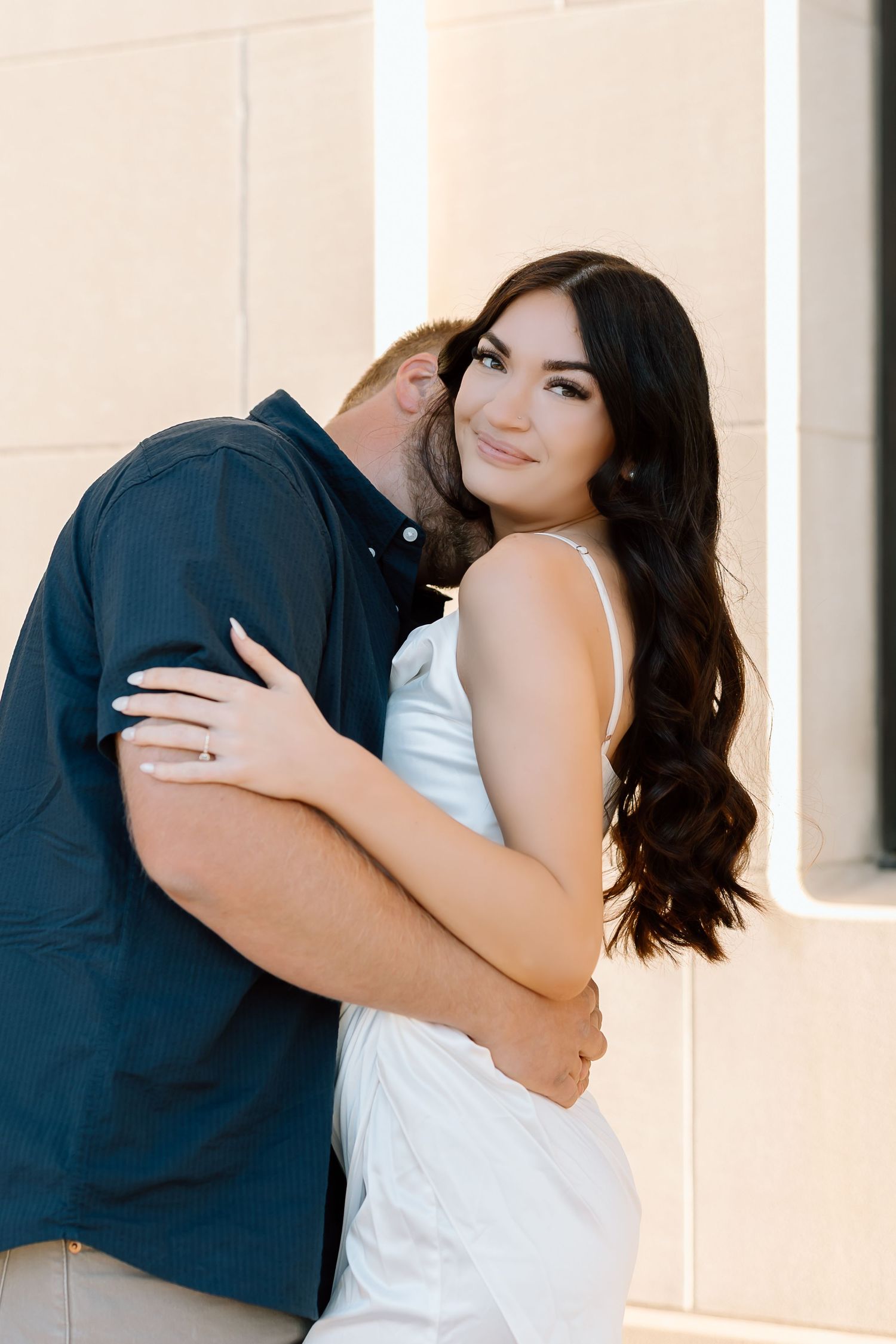 A romantic couple in white and navy blue embraces in a series of intimate poses against a light colored wall.