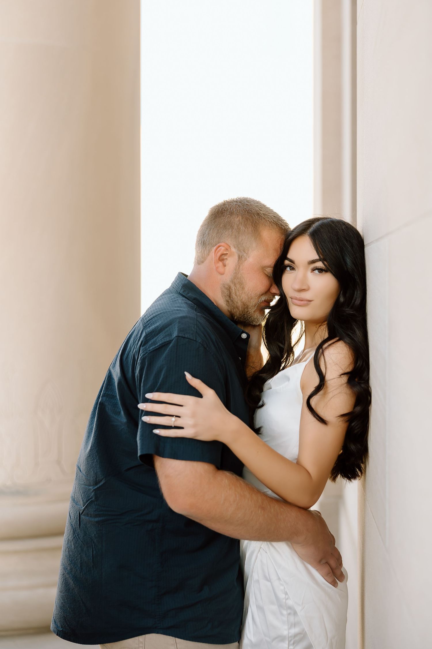 A romantic couple embraces against a light-colored wall, with her in a white dress and him in a navy shirt.