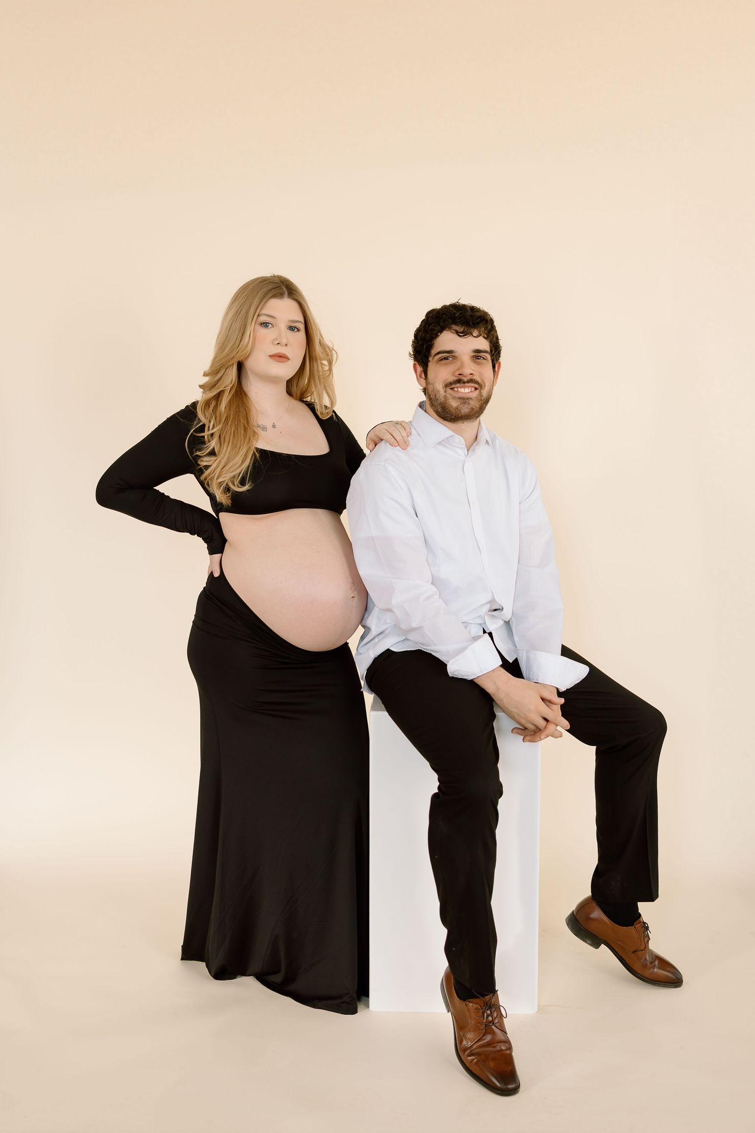 A couple poses for a maternity photo against a beige backdrop, with the woman in black and the man in white.