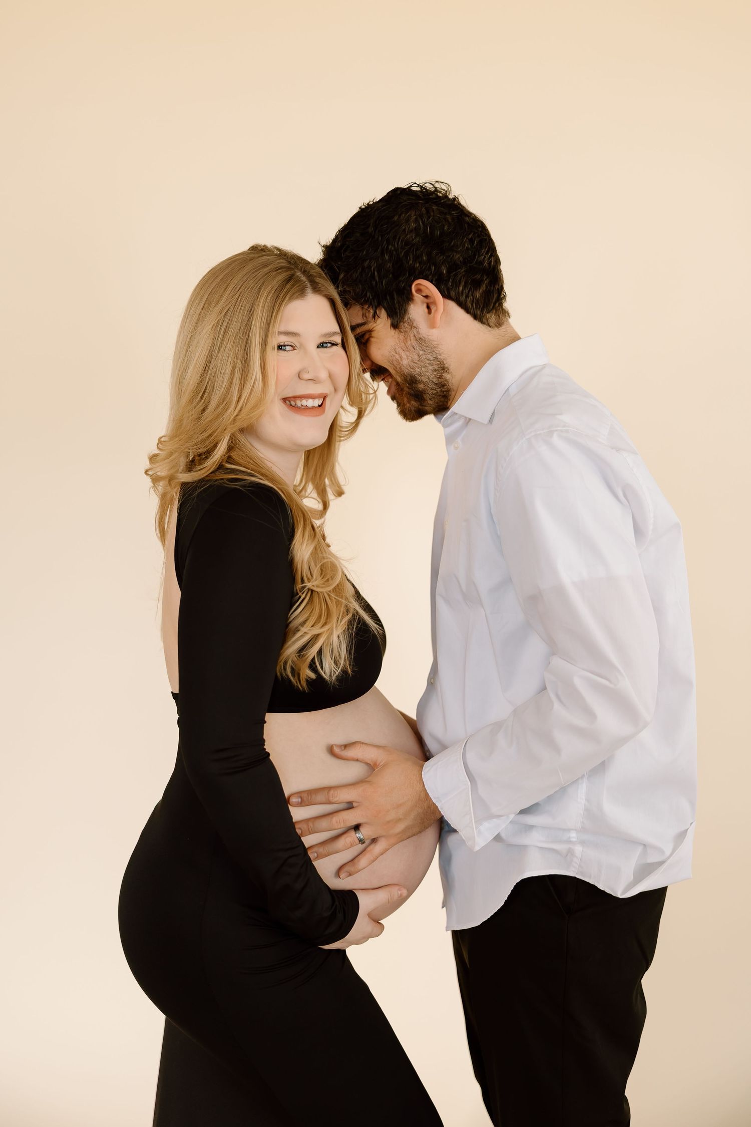 A couple shares an intimate moment during a maternity photoshoot against a neutral backdrop.