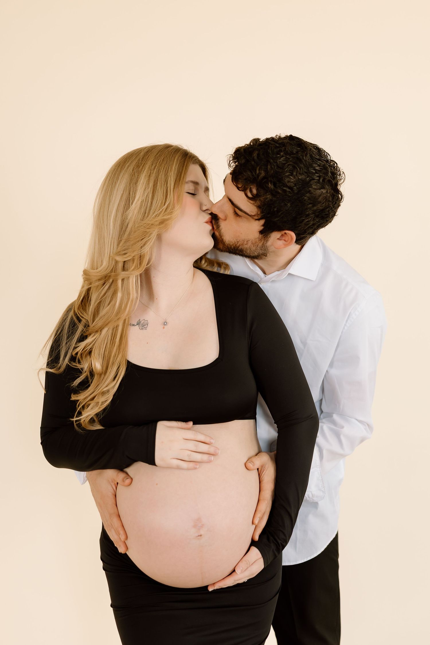 Intimate maternity photo of couple embracing against beige background, with woman in black dress showing pregnant belly.