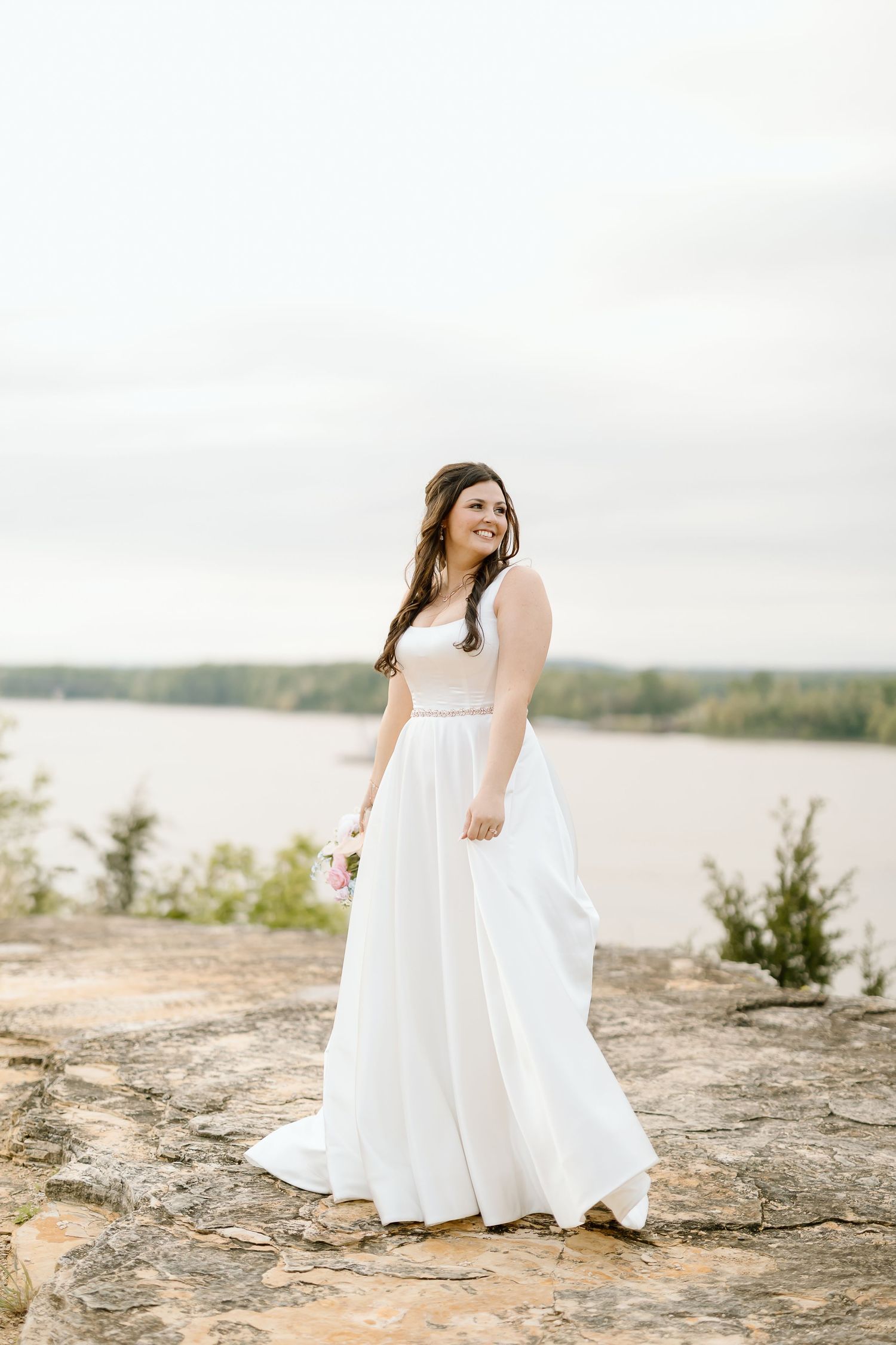 A bride in a flowing white wedding dress poses gracefully on a rocky shoreline overlooking a serene lake.